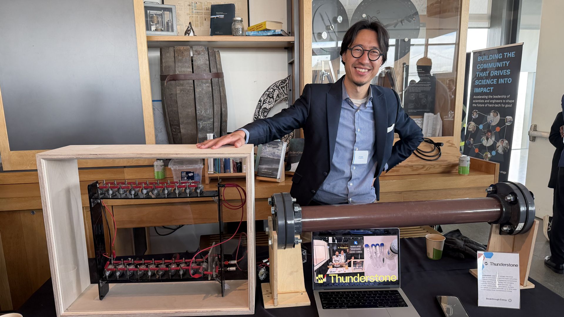 Smiling man with round glasses in a navy blazer leans on a wooden display at a science exhibit. Behind him are shelves with instruments, a large brown cylindrical device, and a Thunderstone banner.
