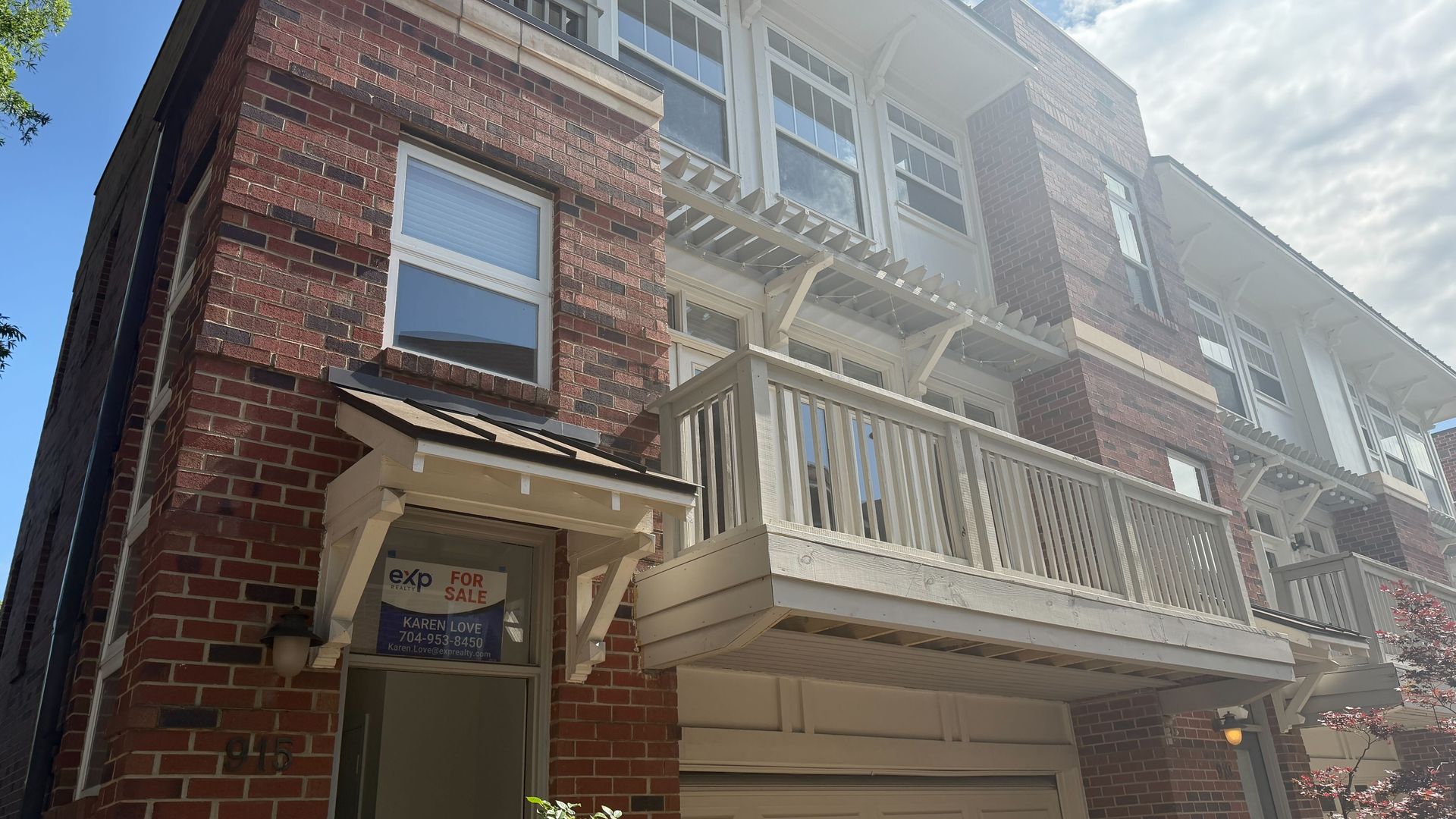 A row of red brick townhouses with white trim, balconies, and a white pergola; a FOR SALE sign visible in a ground-floor window; blue sky with clouds behind.