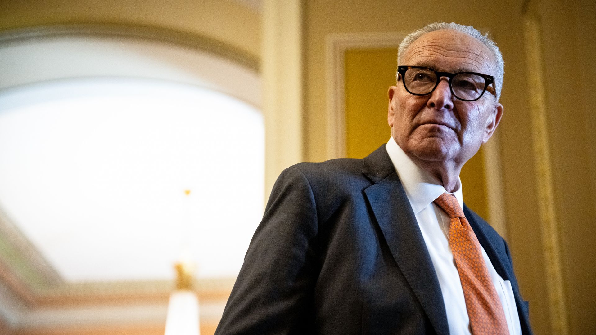 Senate Minority Leader Chuck Schumer, wearing a gray suit and glasses, stands in a hallway.