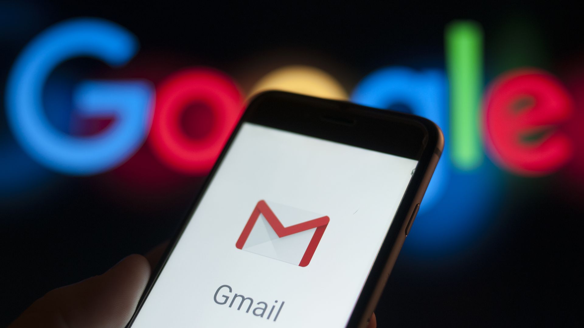 A phone screen with the Gmail app logo on it before the Google logo on a wall.