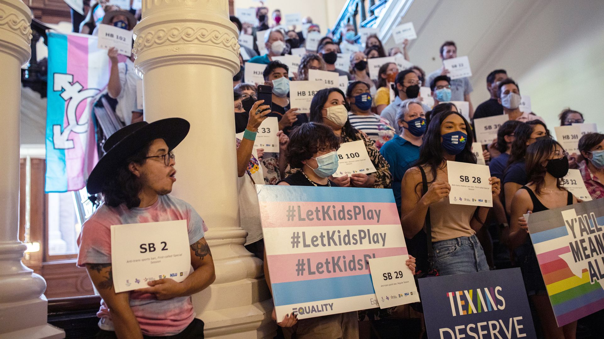 Photo of a crowd of protesters standing and holding signs advocating for trans youth in a building