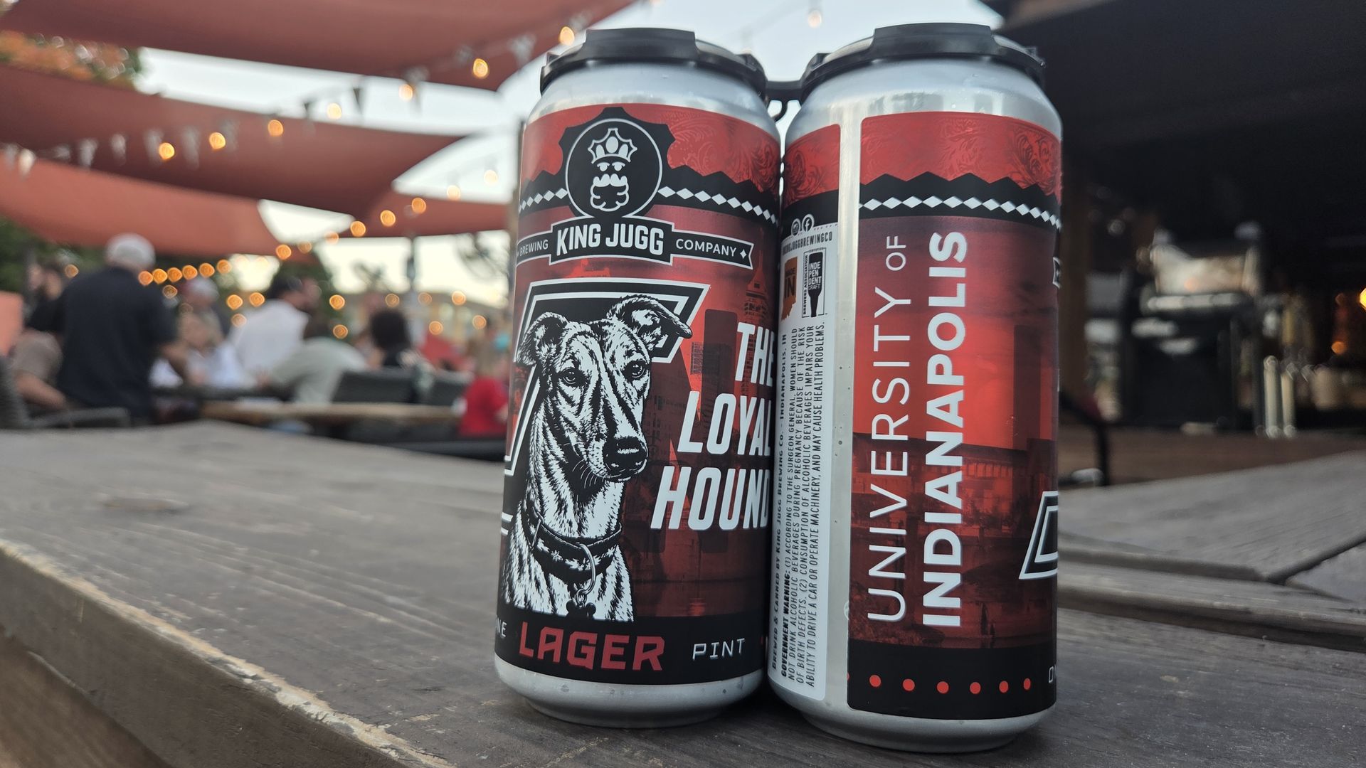 Two red beer cans with a hound illustration and "King Jugg Company" branding sit on a wooden table; blurred outdoor patio with string lights and diners in the background.