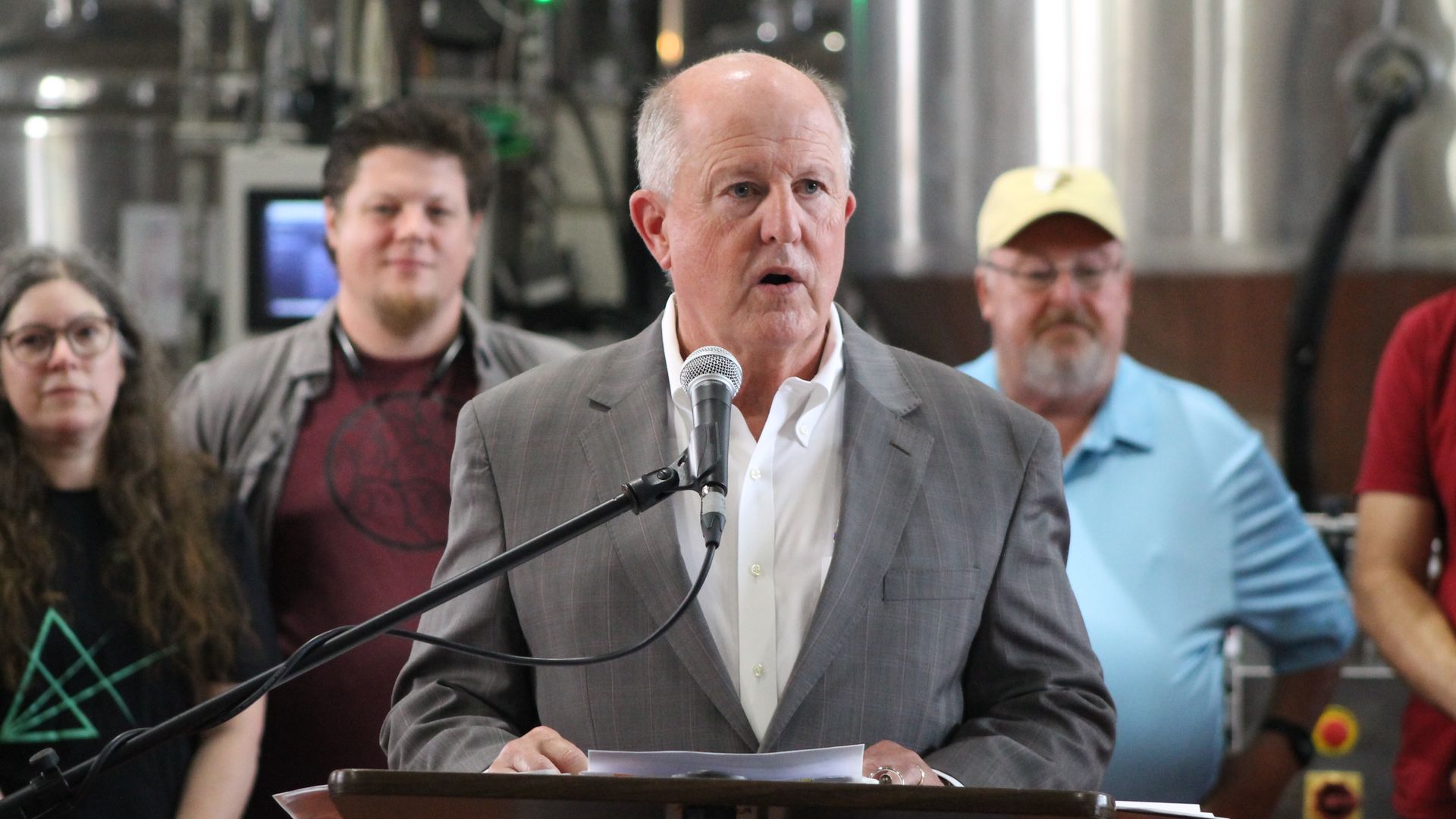 A man in a gray suit and white shirt speaks into a microphone at a podium, with four people standing behind him in a room with industrial equipment.