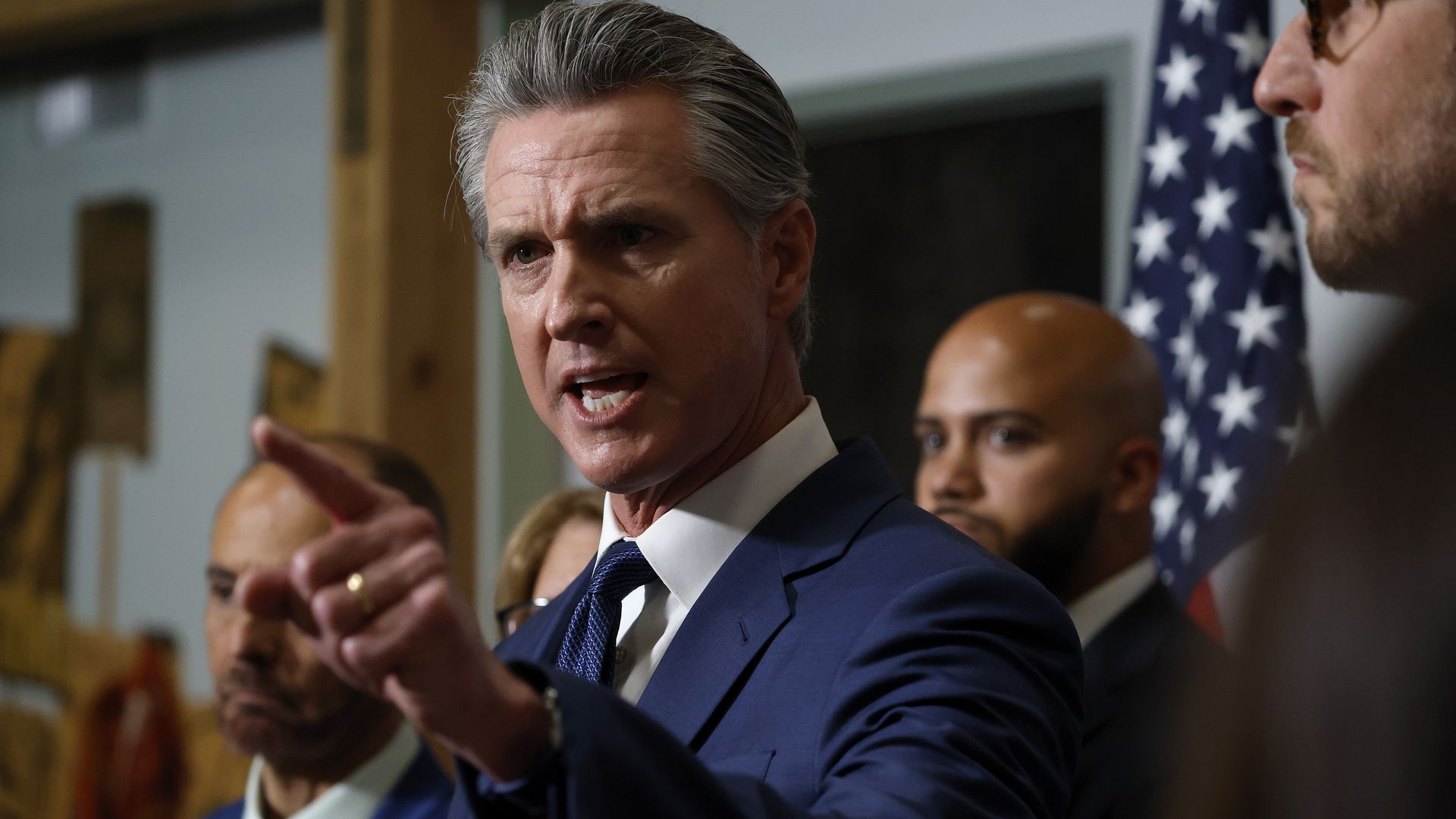 Gov. Gavin Newsom of California in a blue suit and tie speaks emphatically, pointing with his finger. Several people stand behind him, with an American flag visible in the background.