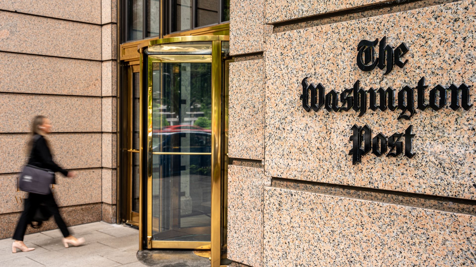 Exterior of the Washington Post headquarters in Washington, D.C., with the newspaper’s name on the stone facade and a person walking past the entrance.