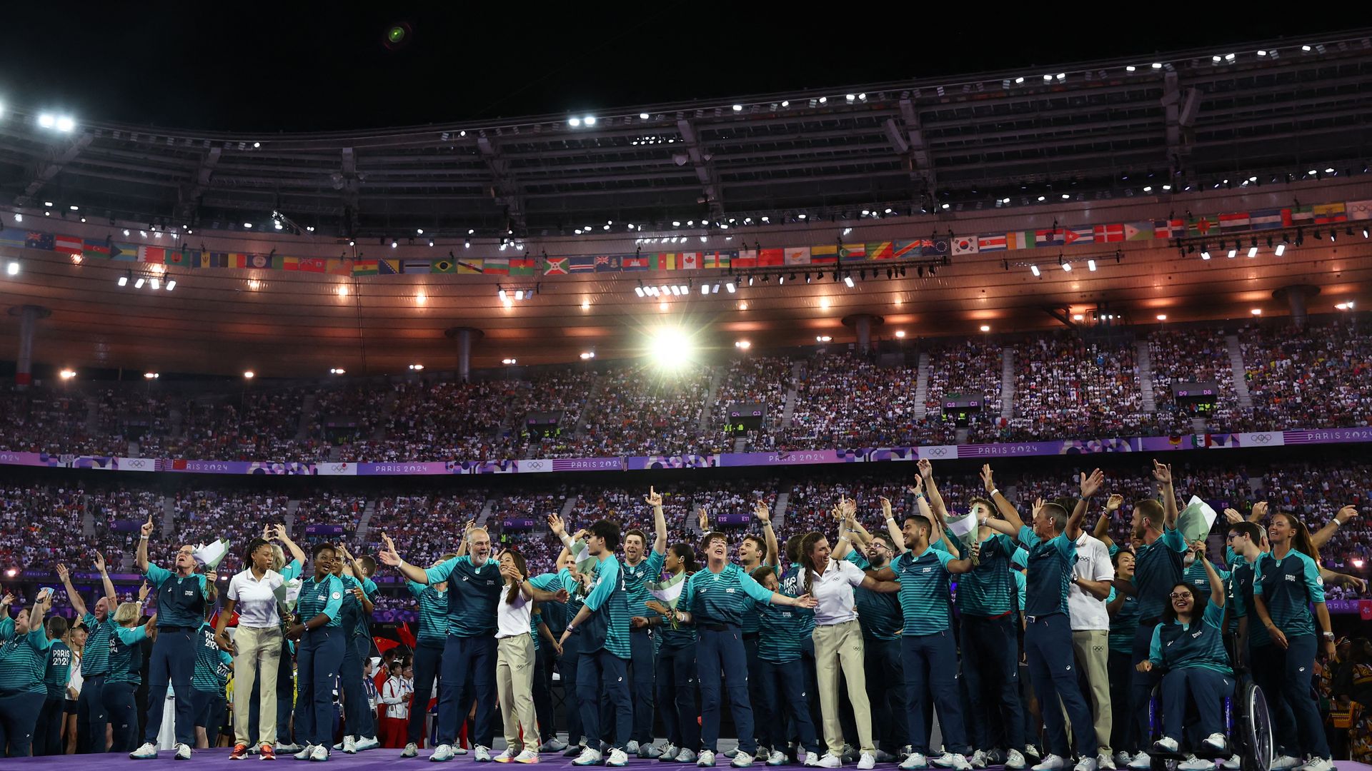Night stadium crowded with fans as Olympic volunteers in teal uniforms stand in a row at the front, hands raised in celebration on stage. International flags line upper railing and Paris 2024 banners appear in arena.