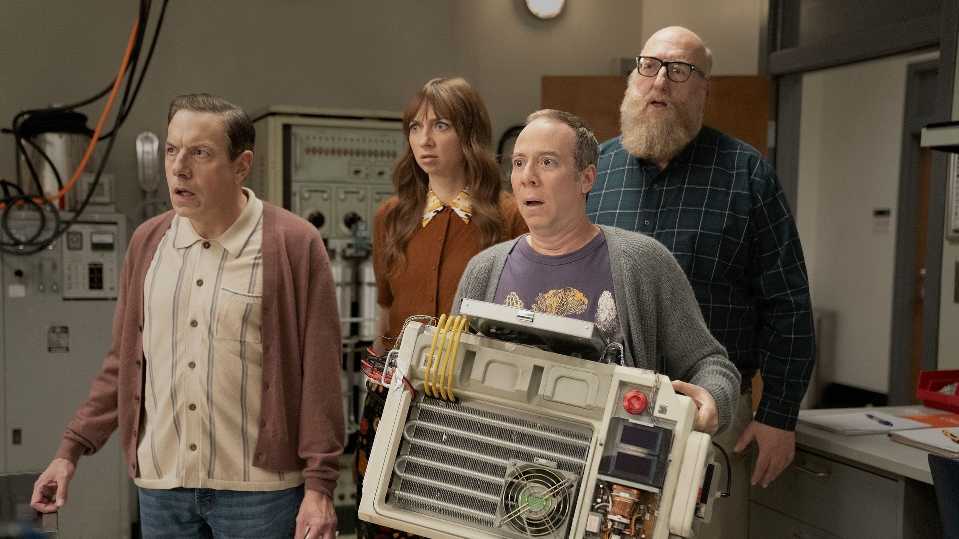 Four adults in a lab look surprised as they carry a large open electronics unit with exposed boards and yellow cables. Background shows control panels and equipment.