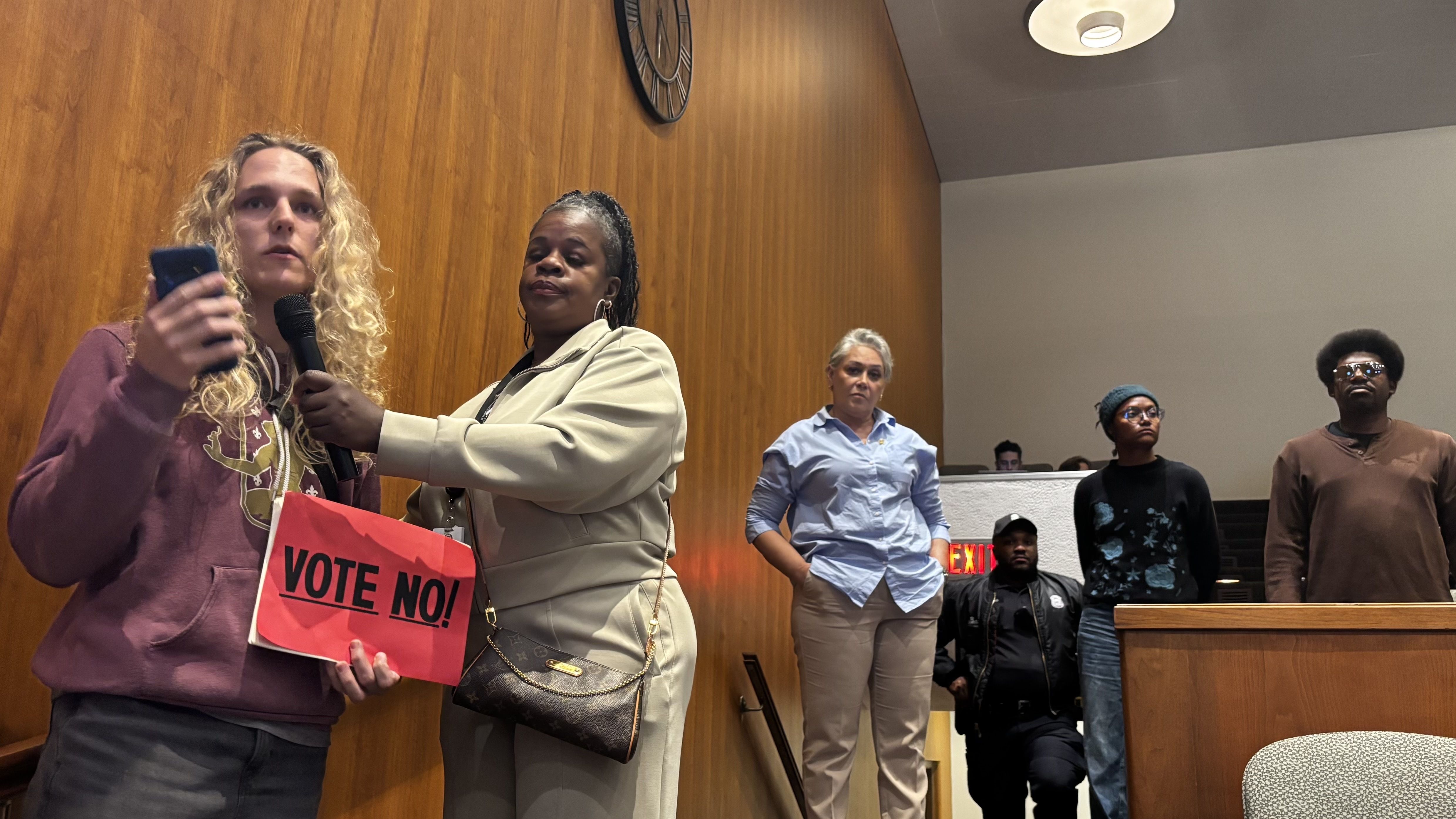 Two people stand at the front of a wood-paneled room; a blonde person holds a pink VOTE NO! sign, while another woman in a beige jacket assists. Several people stand in the background.