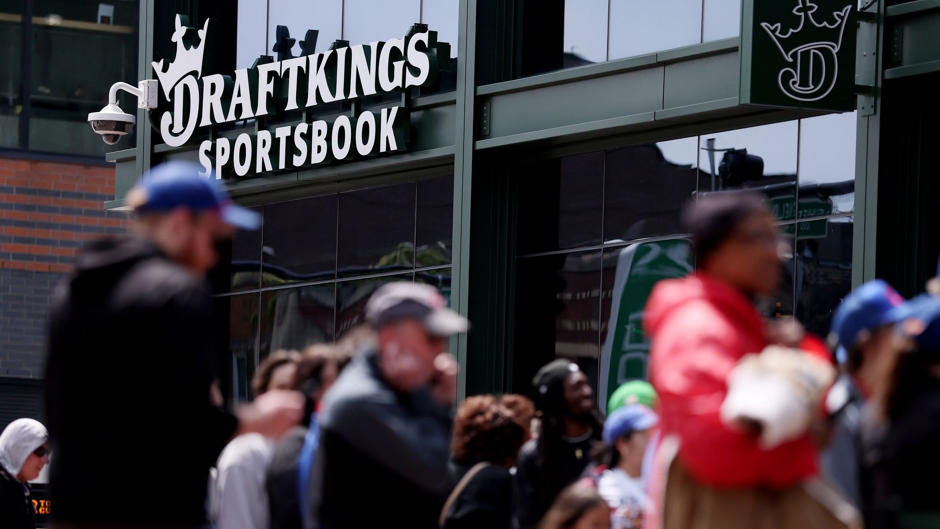 Exterior of a DraftKings Sportsbook storefront with bold white letters and crown logo on glass, a security camera on the left, and blurred pedestrians passing by.