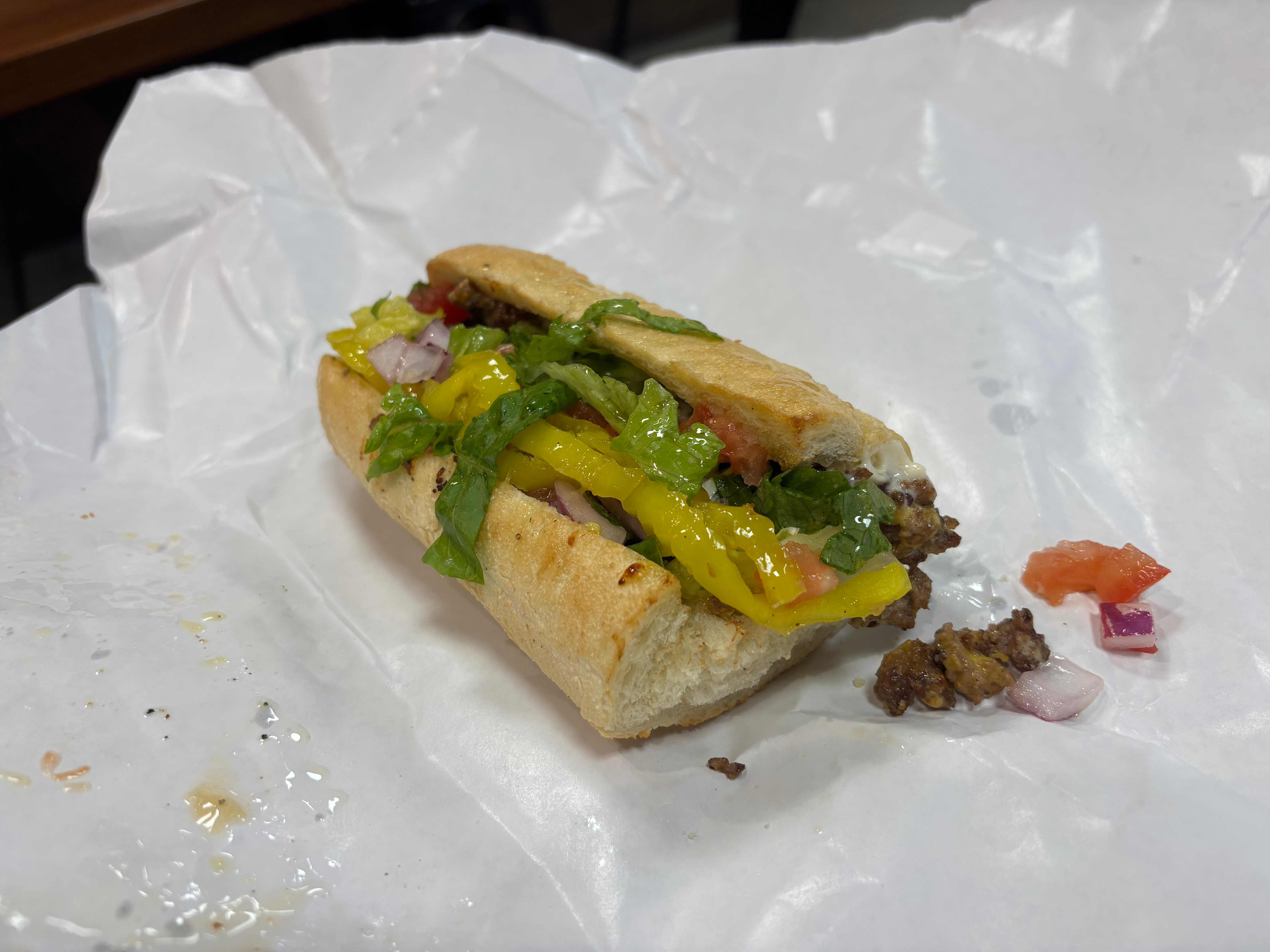 Half-eaten sandwich on a toasted bun filled with yellow pepper slices, chopped red onion, leafy greens, and ground beef, placed on white parchment.