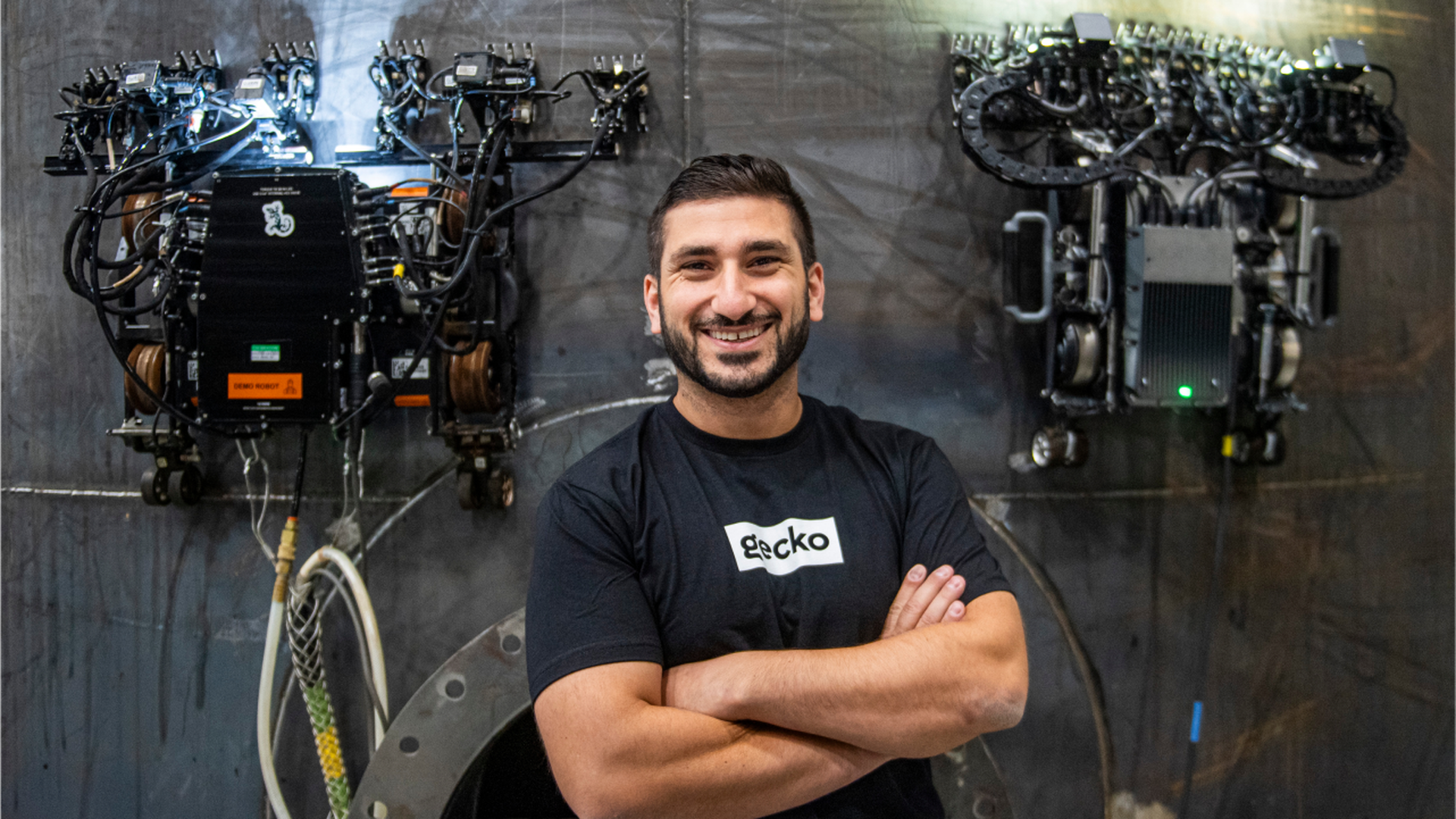 A man stands with his arms folded across his chest. He's in a black shirt. Two robots flank him on the wall behind.