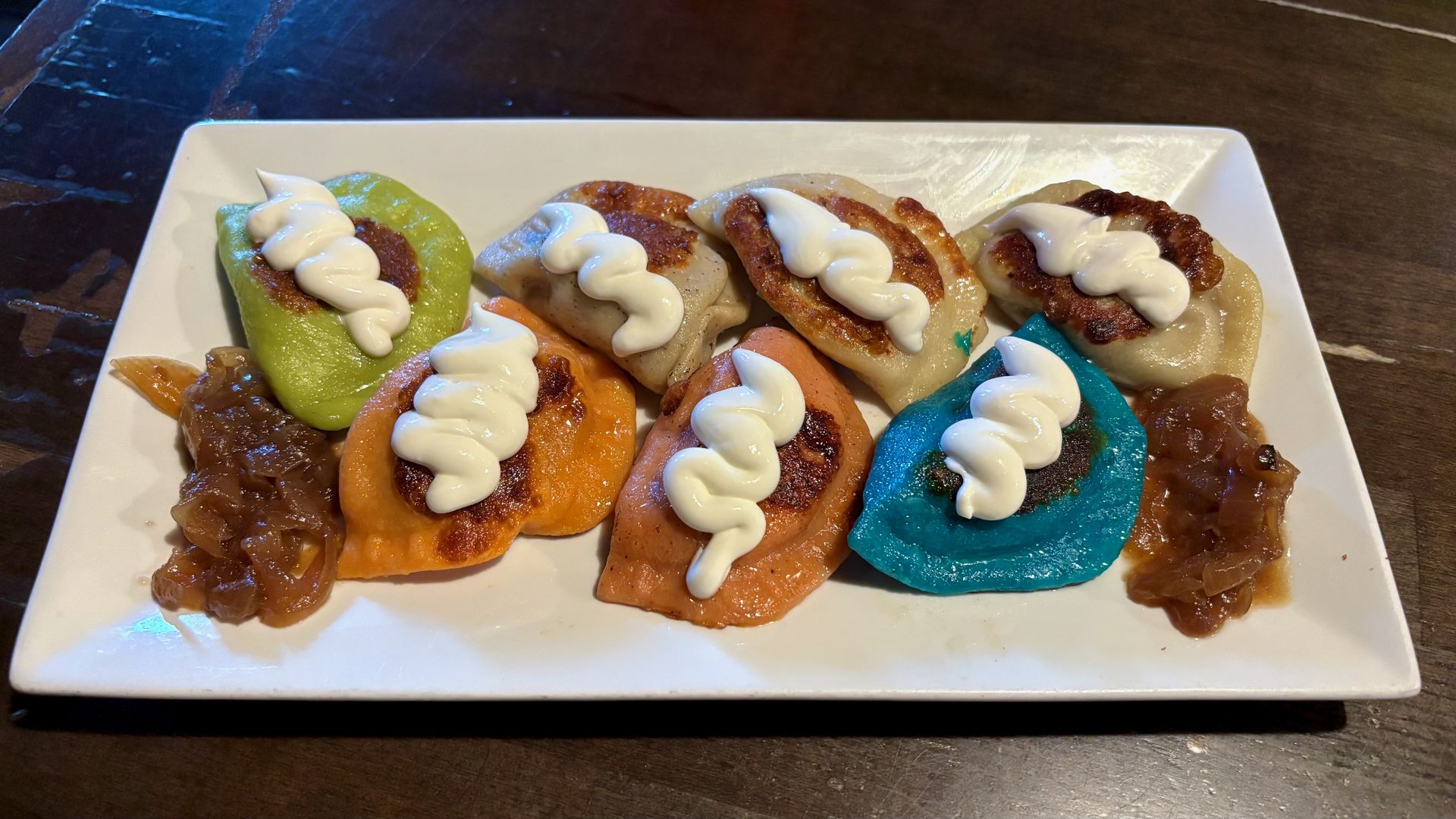 White rectangular plate with several colorful, round pierogi in green, orange, tan, and blue. Each is topped with a white zigzag dollop of sour cream; caramelized onions on the side.
