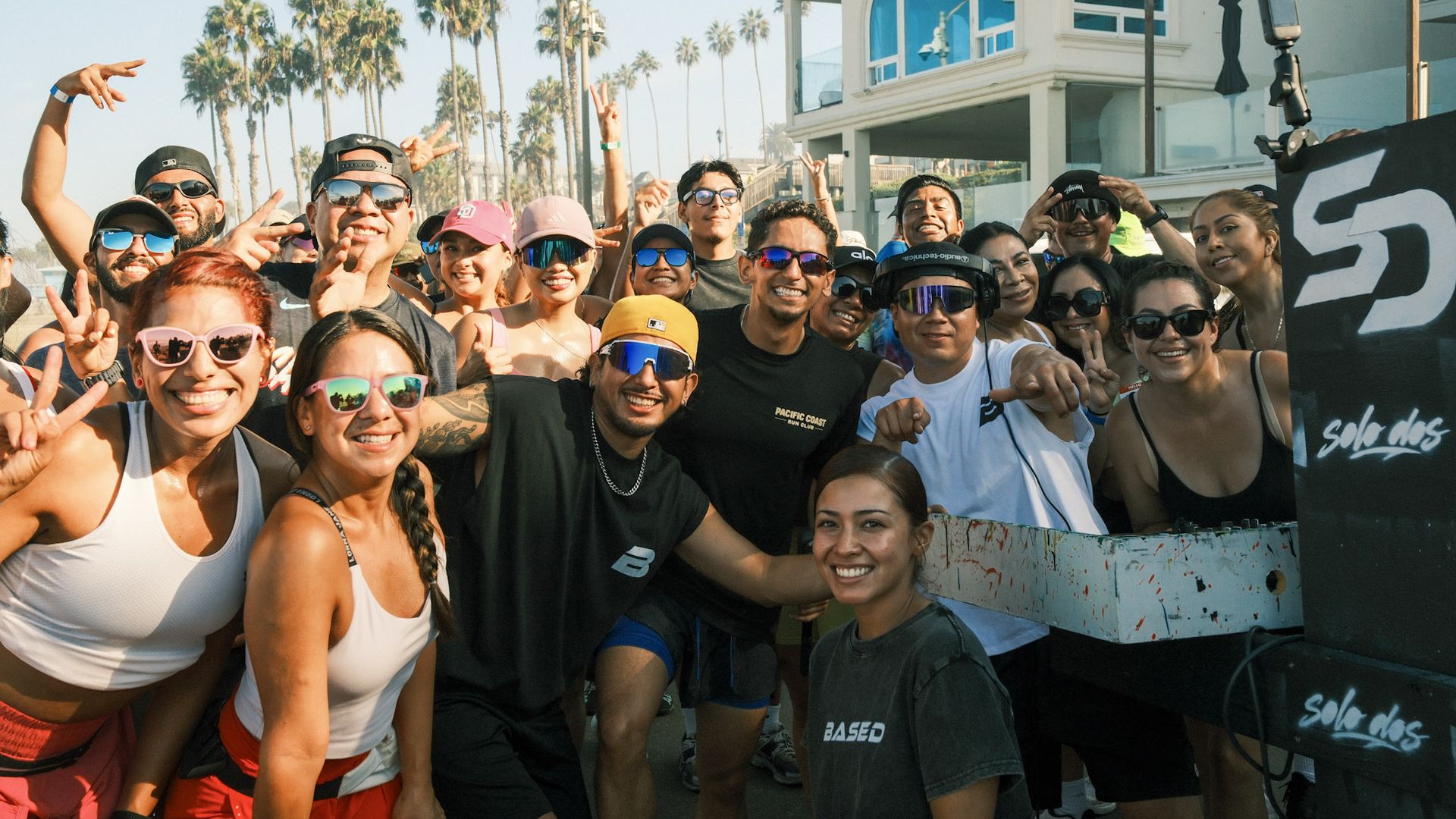 Group of smiling diverse people wearing sunglasses and caps at a sunny beach event, posing toward the camera with palm trees and a white building in the background.