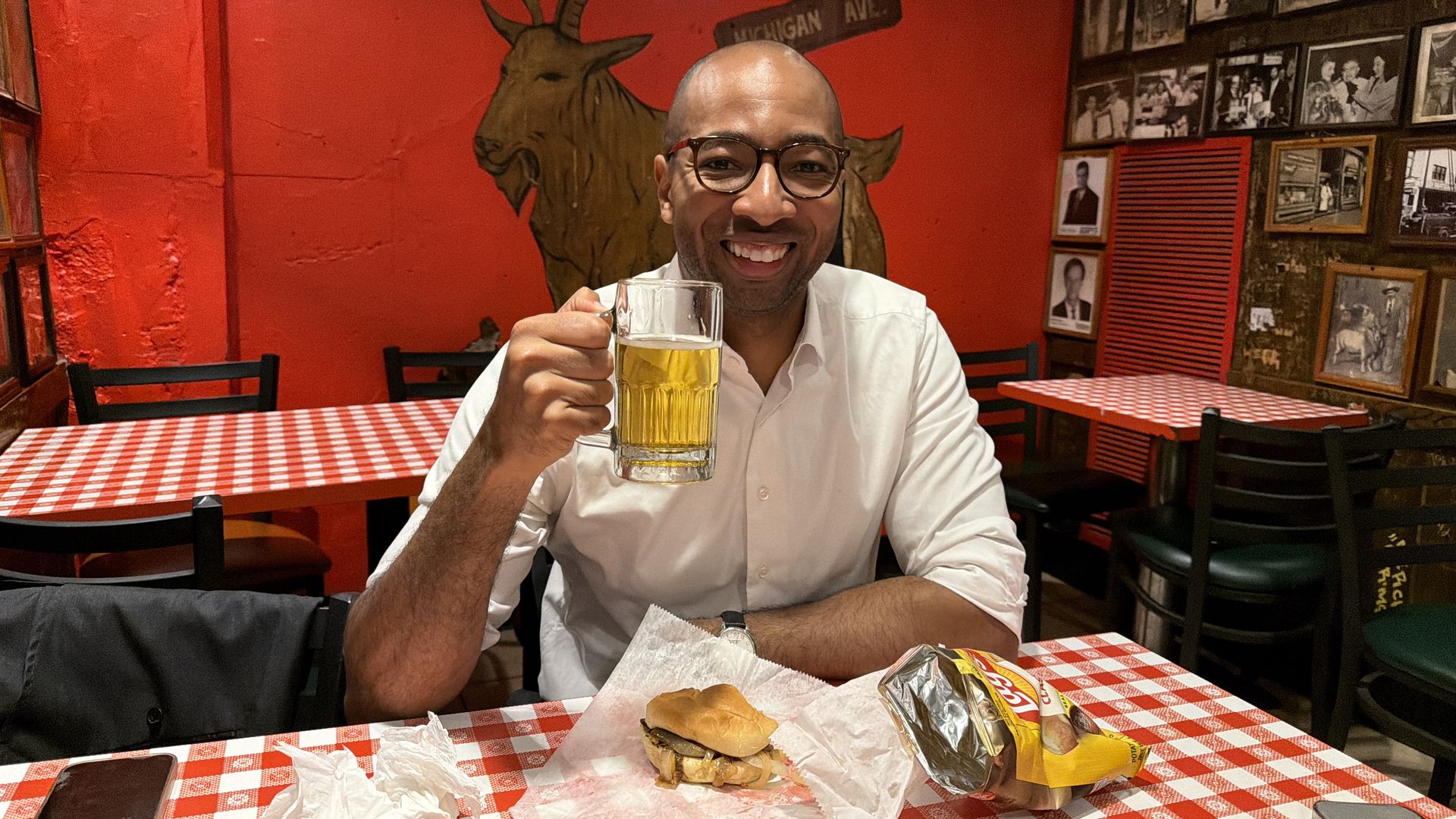 Smiling man with glasses and a shaved head in a white shirt, holding a beer mug at a red-walled restaurant with a goat mural and wall photos; checkered red tablecloth.