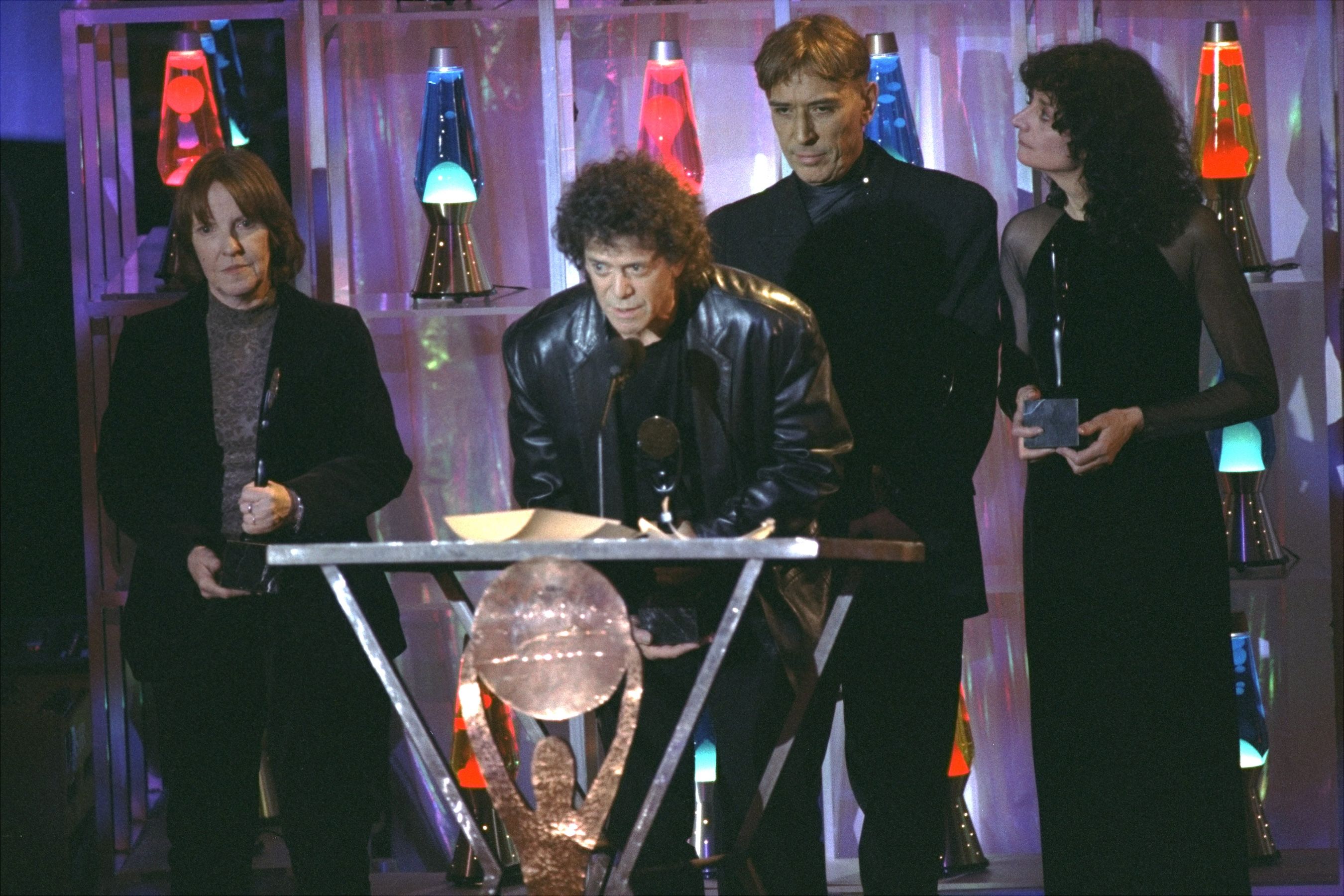 Four people on stage at an award ceremony with colorful lava lamps in the background; one man with curly hair speaks into a microphone at a podium, others hold awards.