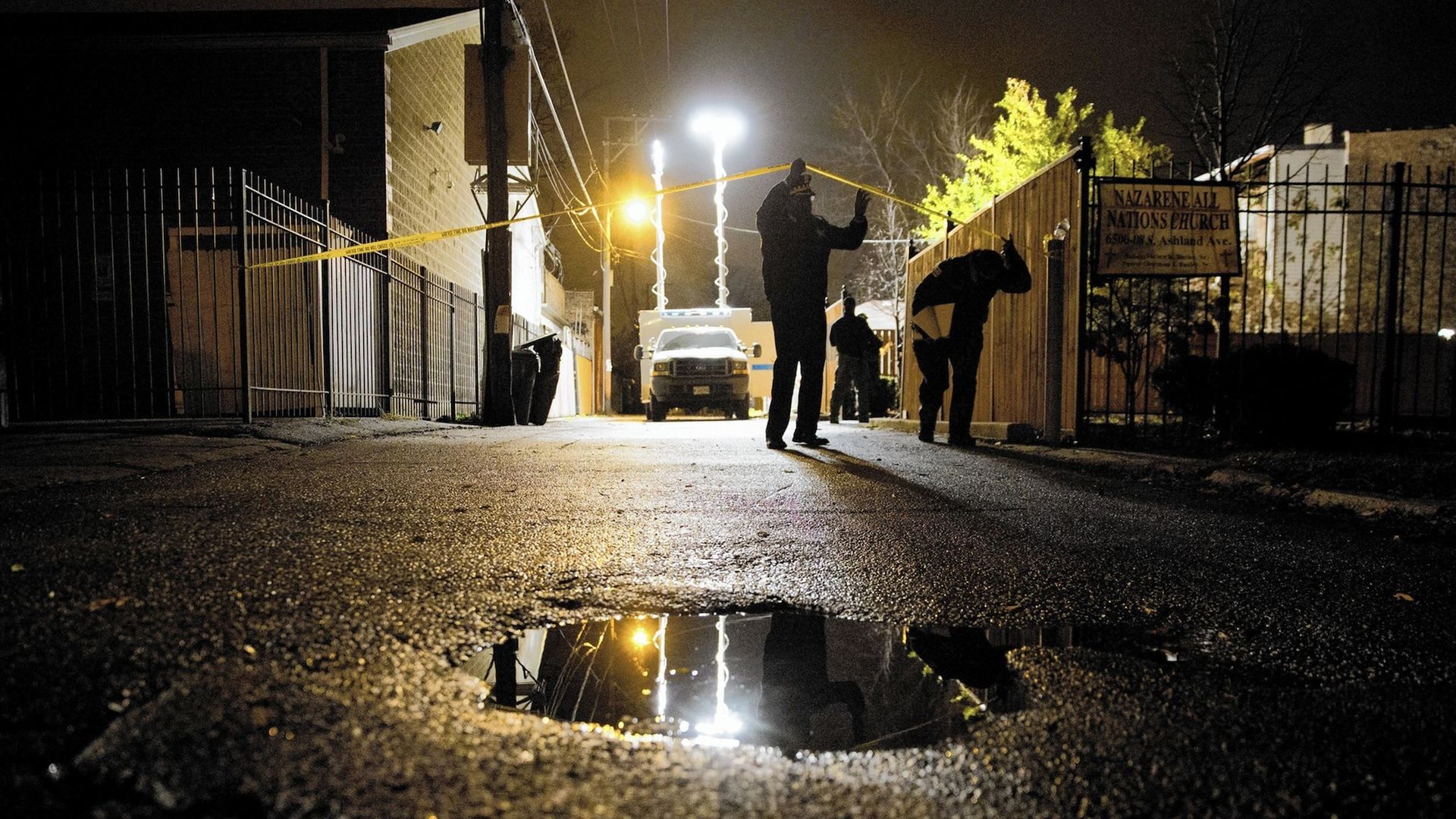 A puddle in a dark alley with a person lifting yellow crime tape.