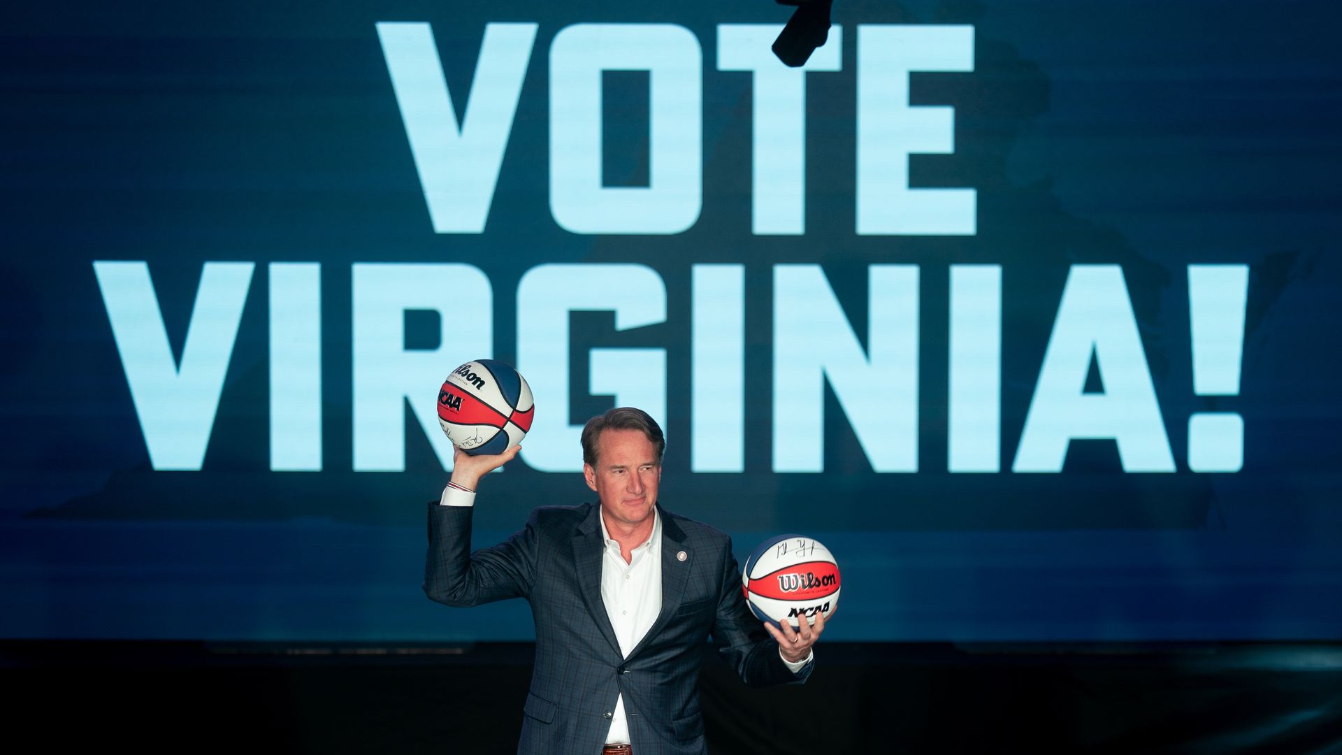 Man in a blue blazer and light pants stands on a stage, holding two basketballs—one in each hand—before a large screen that reads "VOTE VIRGINIA!" in bold white letters.