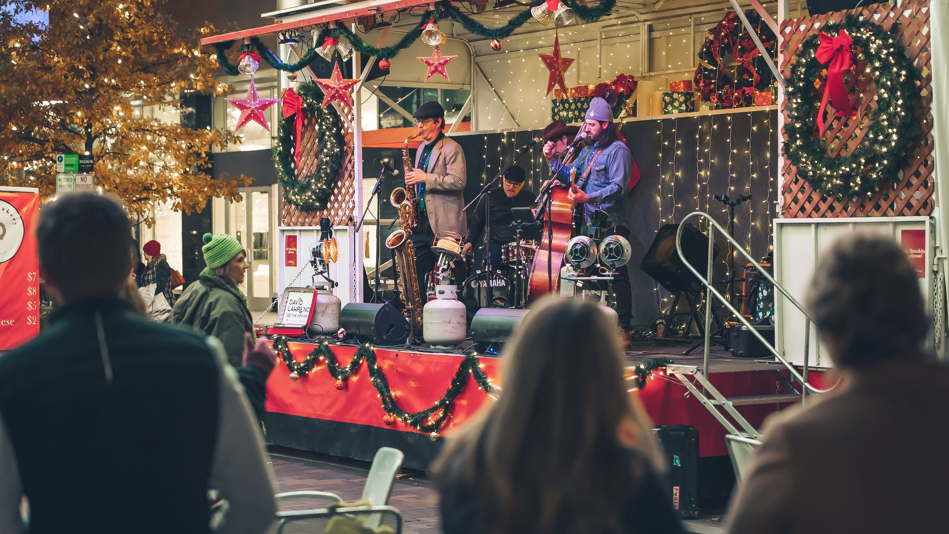 Outdoor Christmas-themed live band performance with musicians playing saxophone, drums, and double bass on a decorated stage with wreaths, red stars, and string lights, audience watching.