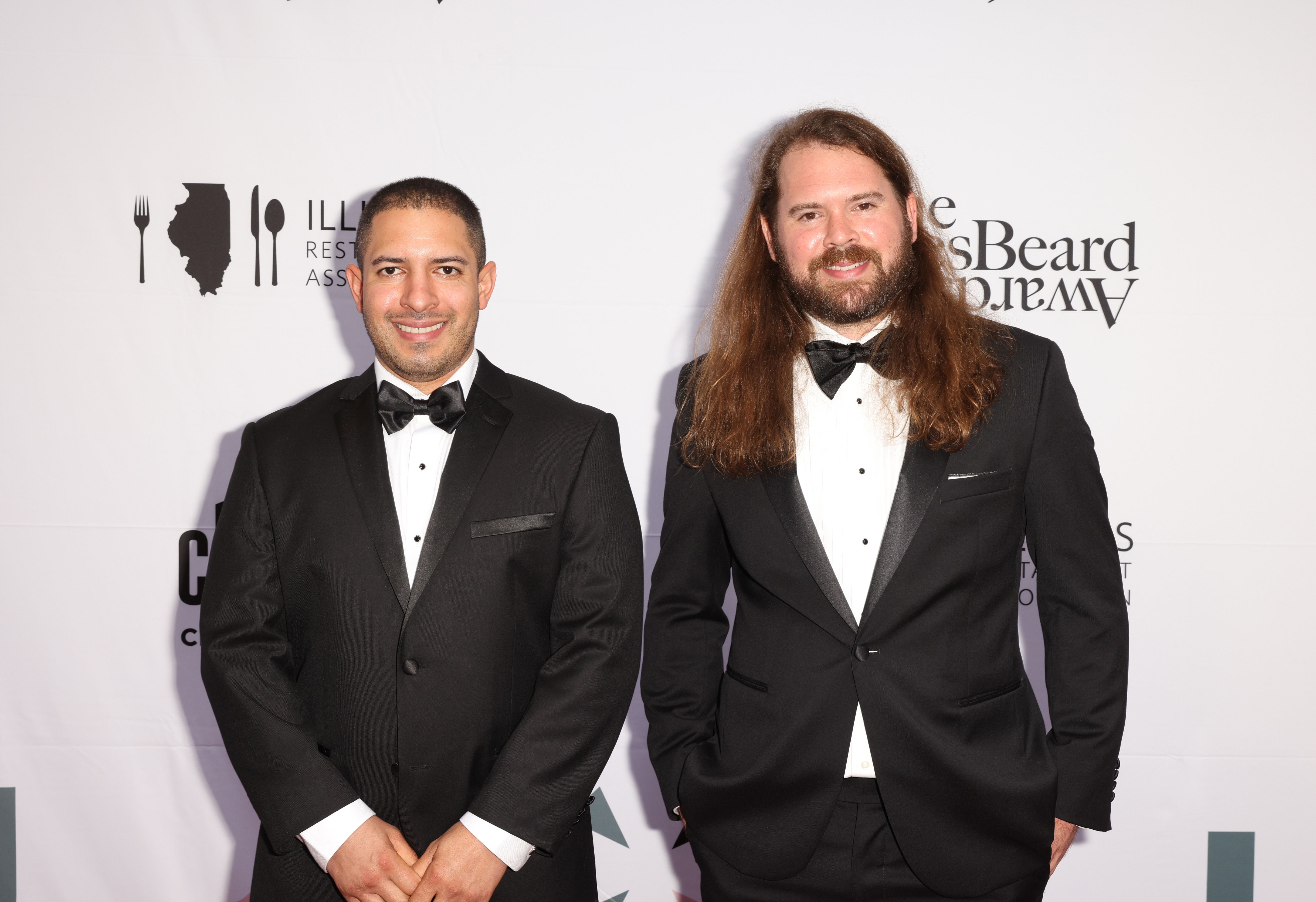Two men wear tuxedoes and smile for a photo.