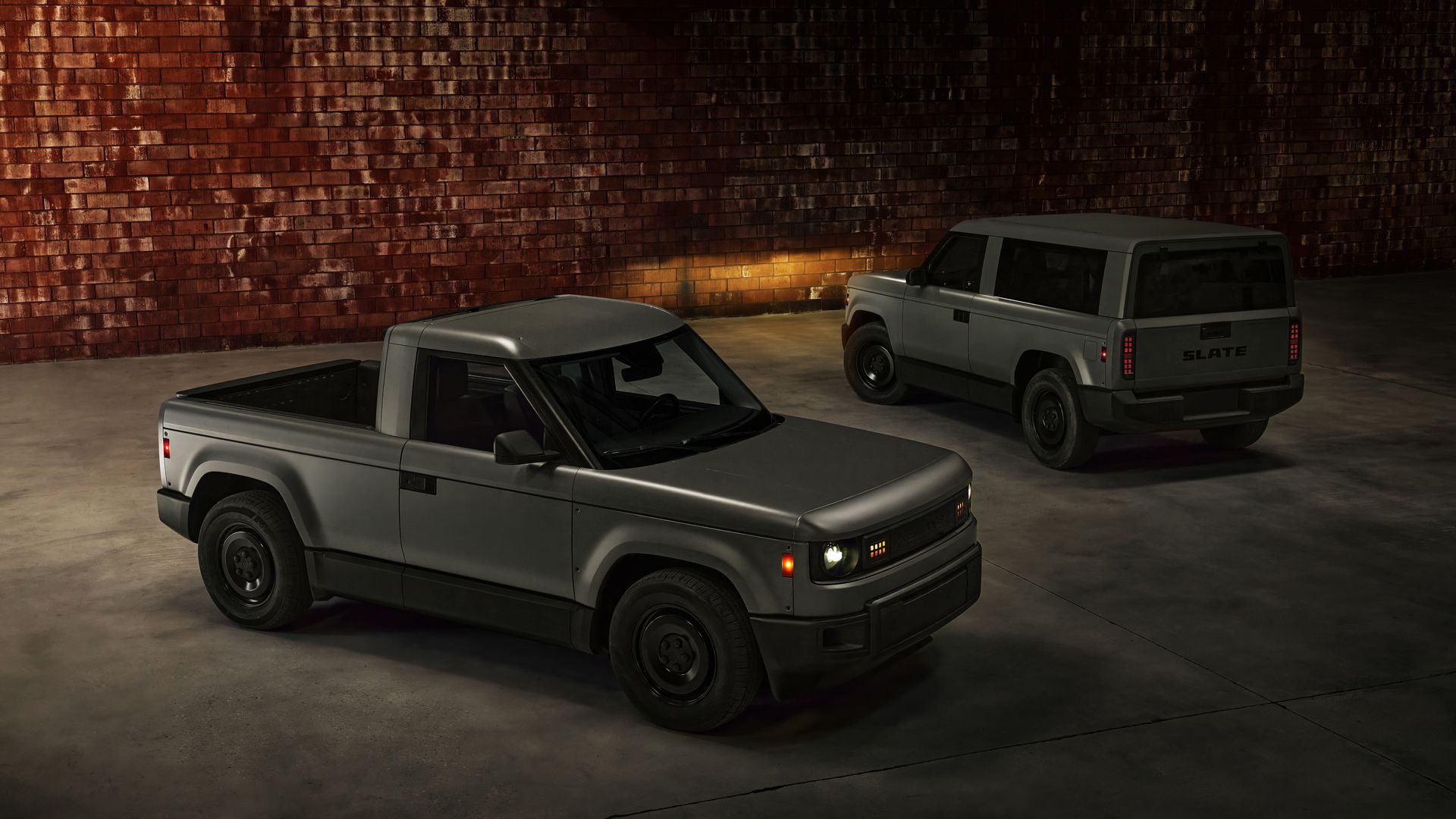 Dim indoor parking area with a red brick wall; three gray vehicles are parked along the wall, including a pickup in the foreground and an SUV with "SLATE" on its tailgate.