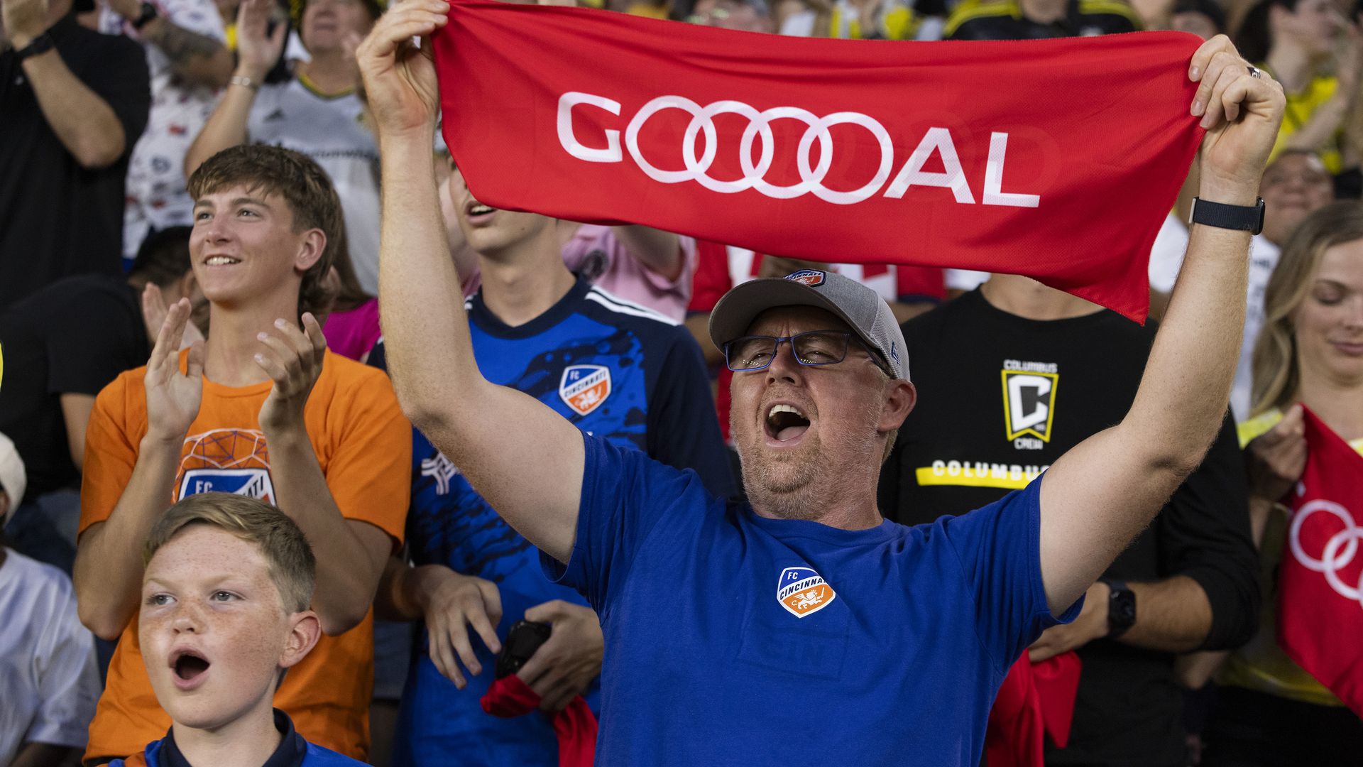 Fans cheer and one holds up a banner reading "GOOOAL" at a soccer match.