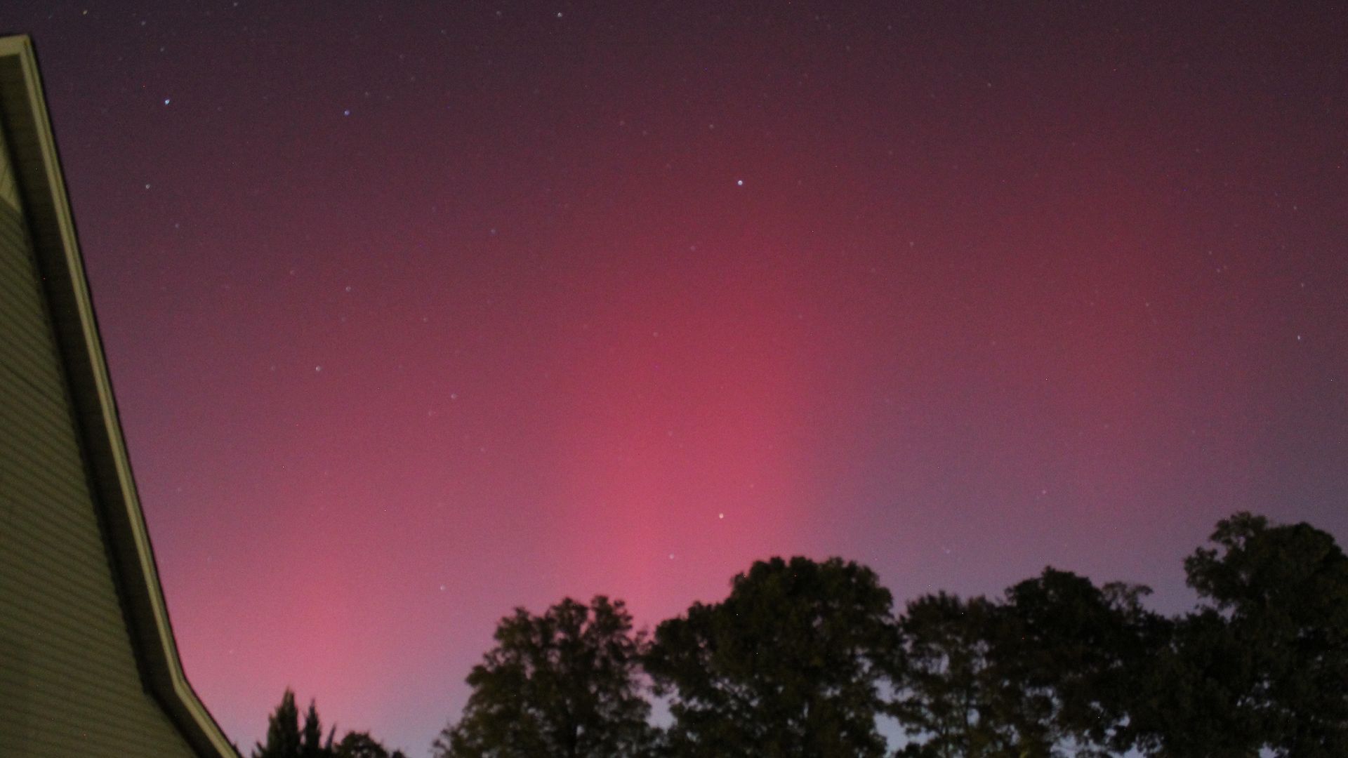 Night sky with red and purple aurora above dark tree silhouettes and a house on the left side, with stars visible throughout the sky.