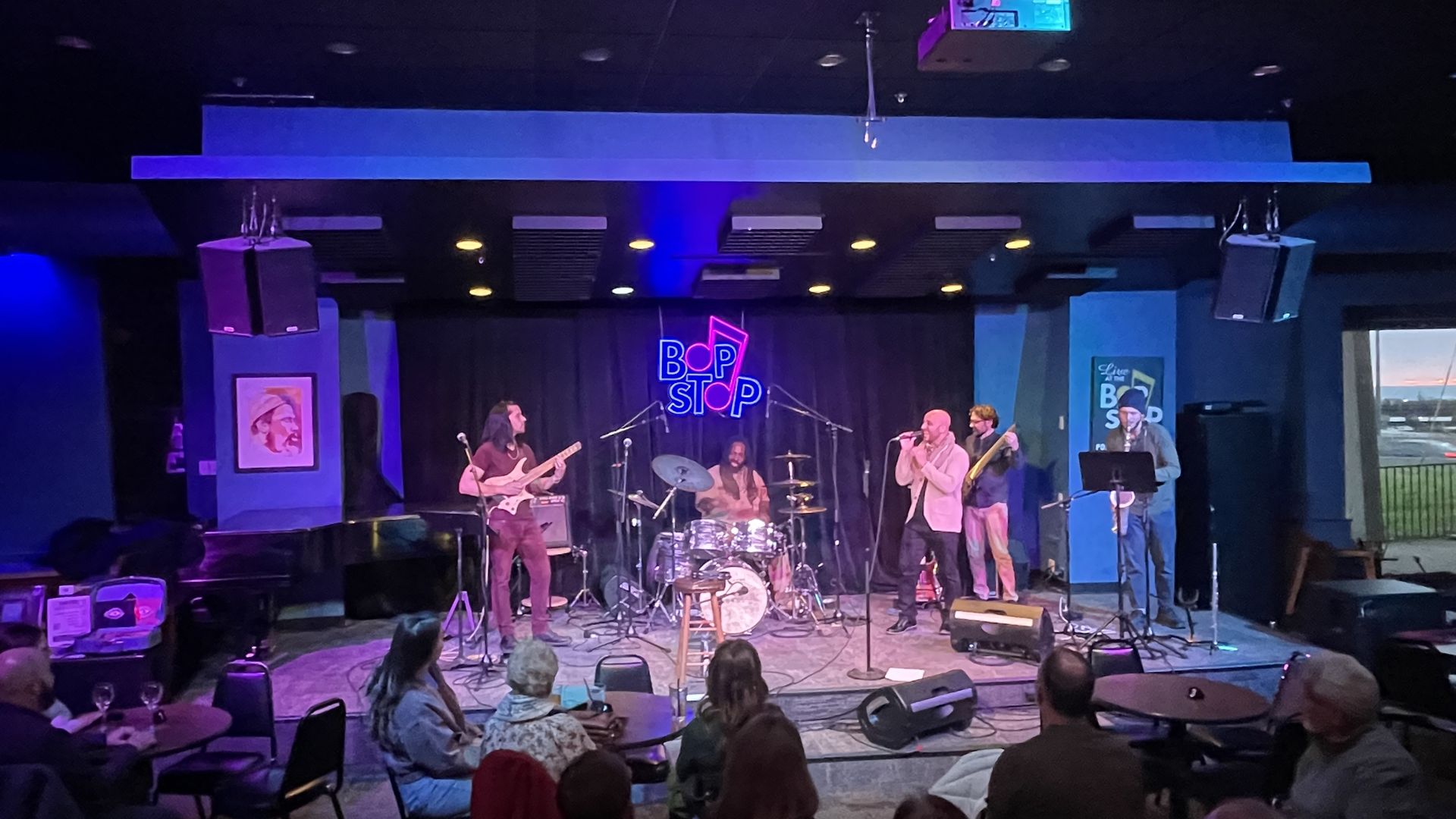 Band on stage at Bop Stop venue with blue and purple lighting. Left guitarist, center drummer, right lead singer with mic, bassist beside. Audience seated at tables in front.