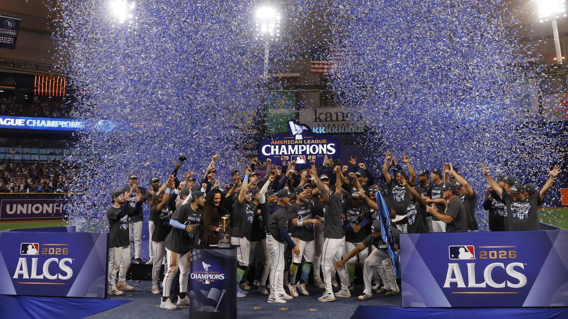 Baseball team on a podium celebrating amid a blue and white confetti shower; cheering fans surround banners reading "American League Champions" and "ALCS 2026," with a trophy at center stage.