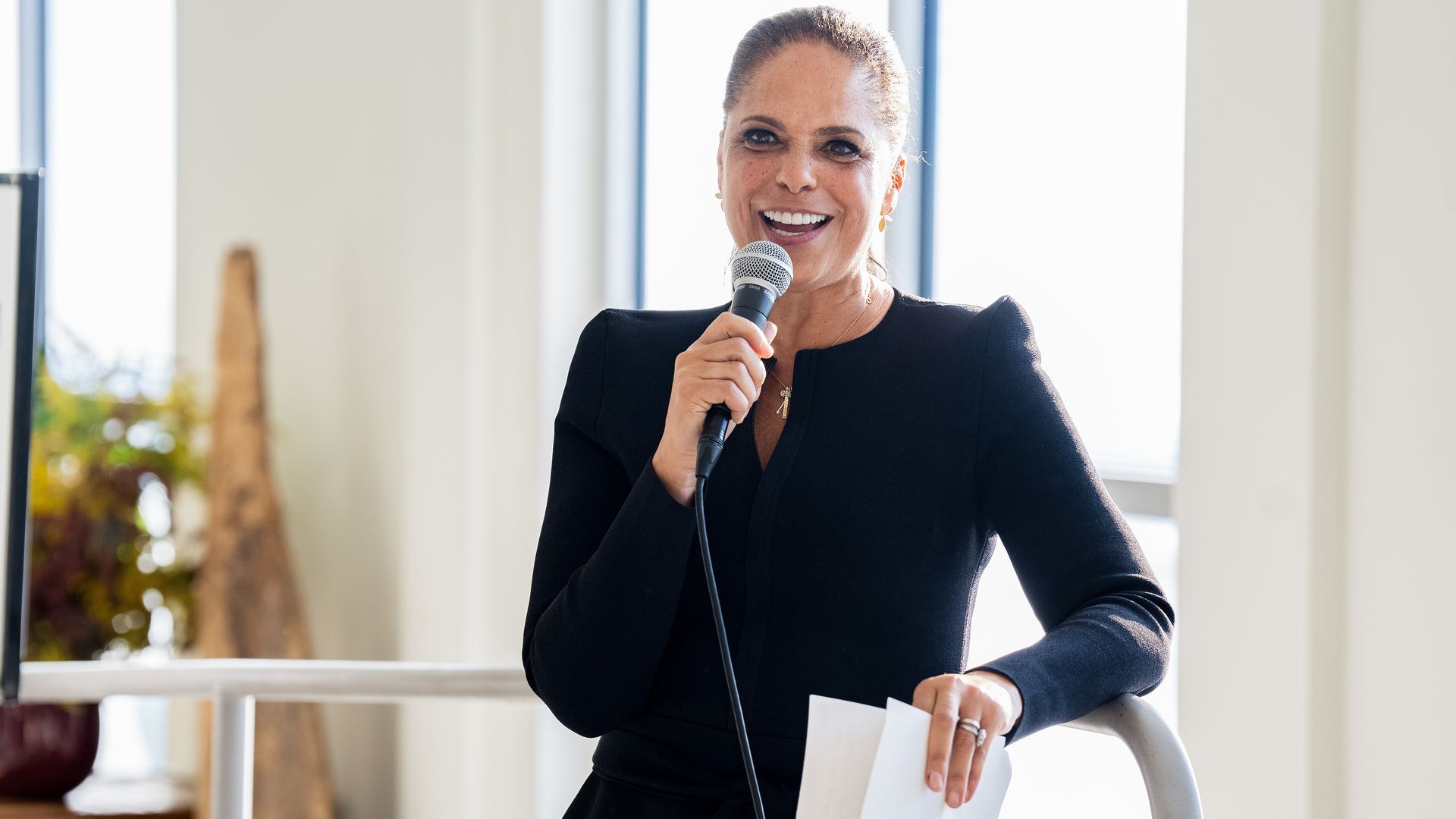 A professionally-dressed woman leans against a railing. She holds a microphone to her mouth in one hand and a couple of papers in the other.