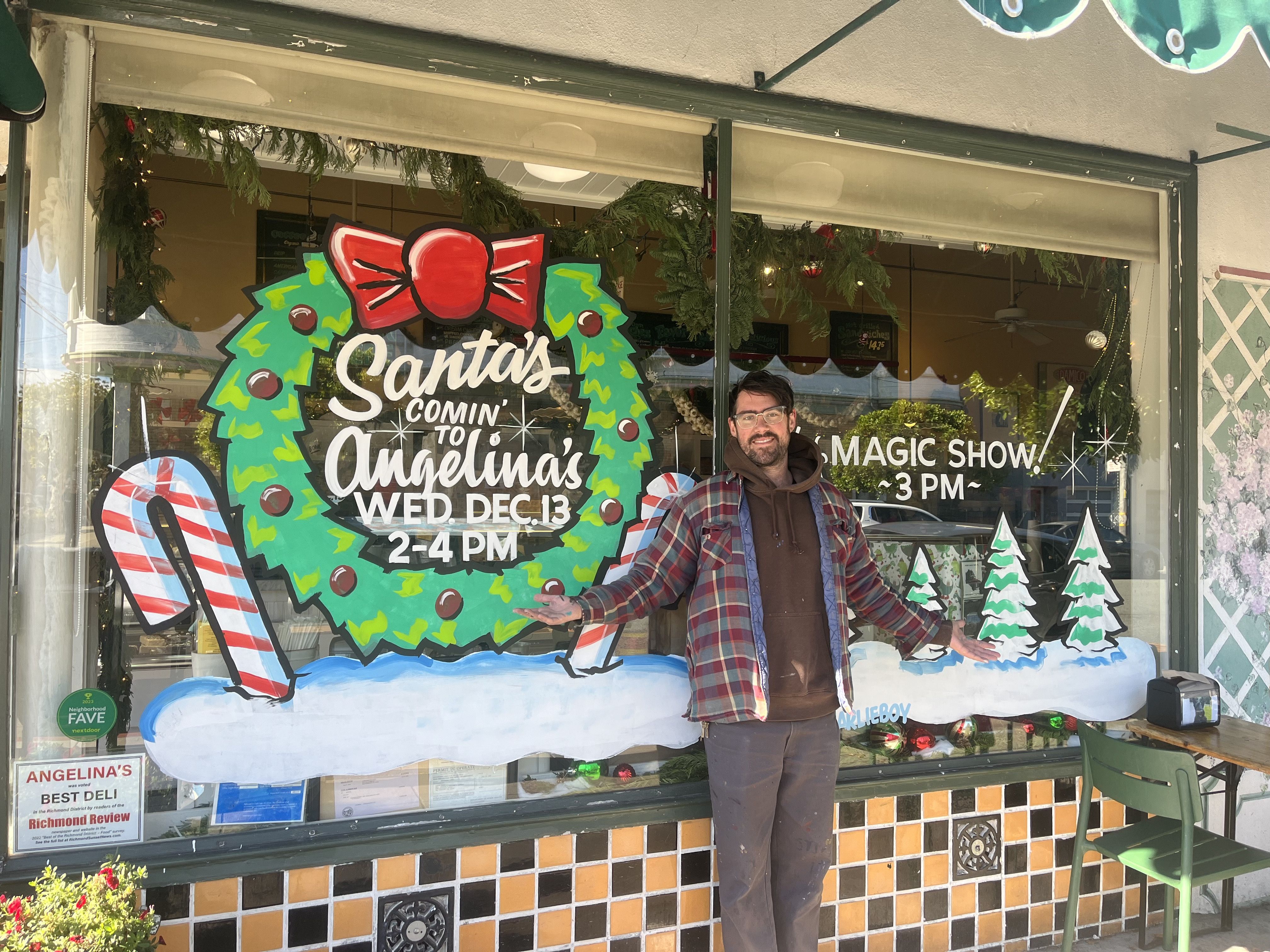 Man in glasses, brown hoodie, and plaid jacket stands smiling in front of shop window decorated with a festive Christmas wreath, candy cane, and snow-covered pine trees advertising Santa visit and magic show.