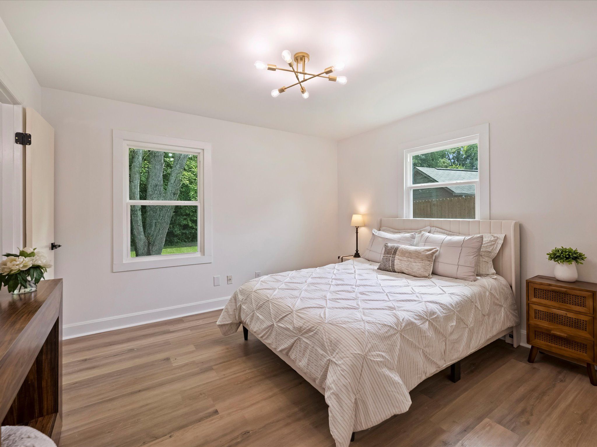 Bright bedroom with white walls and wood floor, featuring a bed with white and beige bedding, two windows showing greenery outside, a wooden dresser, nightstand, lamp, and small plants.