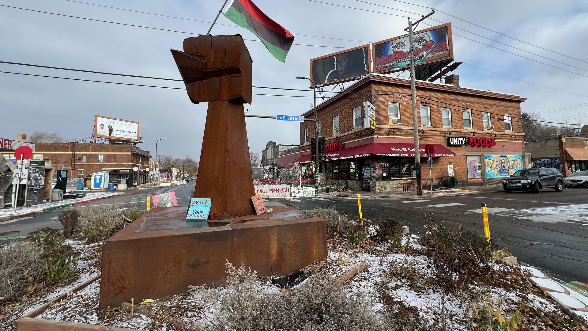 Massive rusty metal fist sculpture on a round pedestal, waving a red, black, and green flag; an urban street with a brick building labeled Unity Foods, billboards, and light snow.