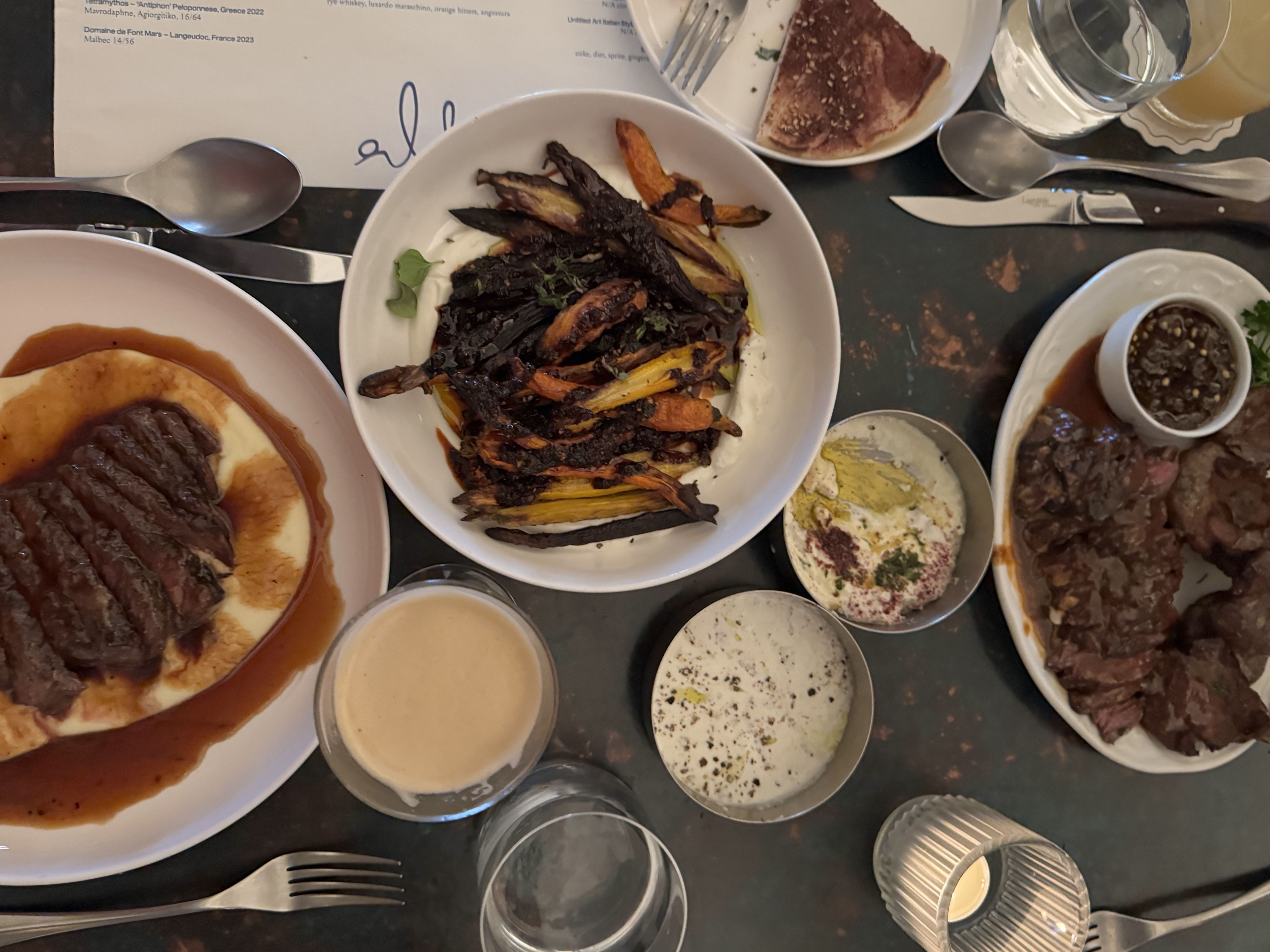 A table setting featuring a variety of dishes. On the left, a plate with sliced meat in sauce on mashed potatoes. In the center, a bowl of roasted carrots and other vegetables. On the right, a plate with steak and a small bowl of sauce. 