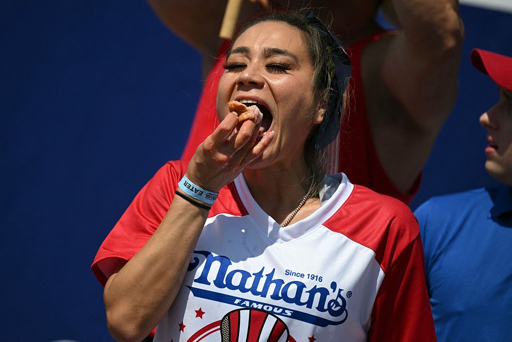 A woman wearing a red, white and blue Nathan's shirt putting a hot dog with bun in her mouth.