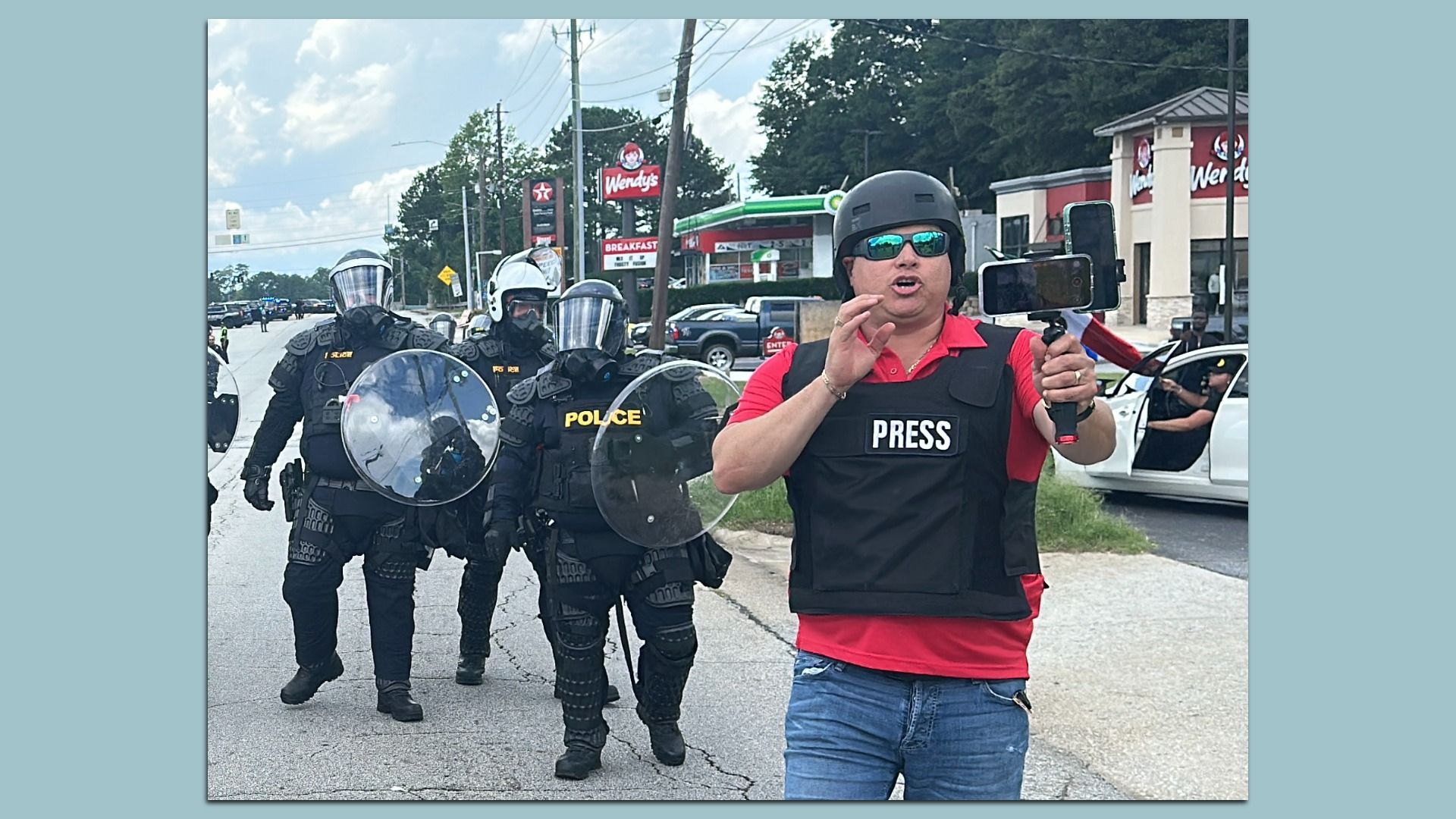 A man wearing blue jeans, black helmet, sunglasses, a black vest with the words 'PRESS' over a red shirt films himself as he walks along the street with three police officers dressed in riot gear follow.