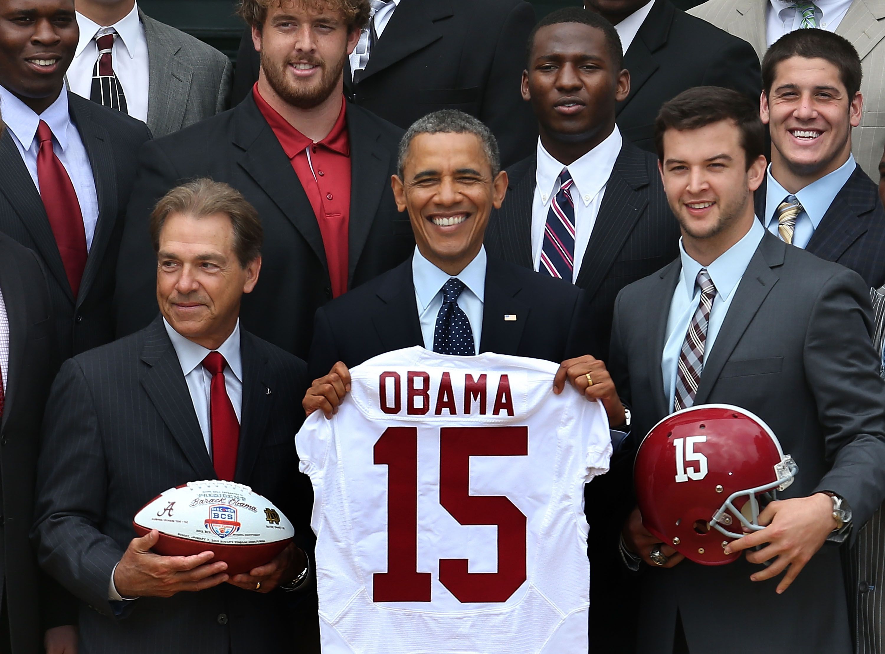 Picture of Barack Obama surrounded by members of the Alabama football team while holding a football jersey that says "Obama" on the back with the number 15