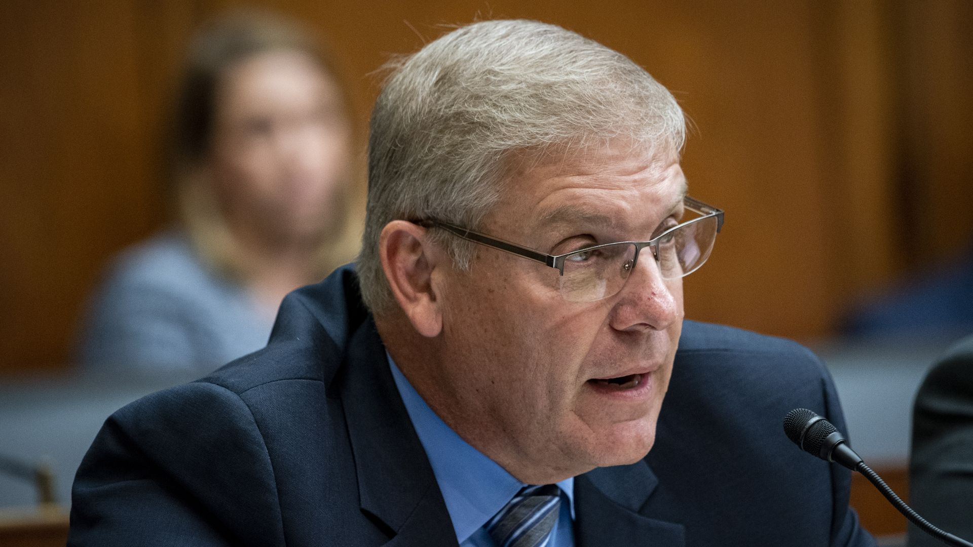 Rep. Barry Loudermilk, wearing a dark blue suit jacket, medium blue shirt, blue and white striped tie and glasses, speaking into a microphone.