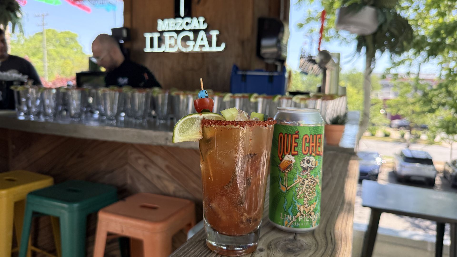 Two cocktails on a wooden bar with lime wedges and a blue skewer topper; a green skeleton-themed can beside. Blurred background shows a bar and a neon sign reading MEZCAL ILLEGAL.