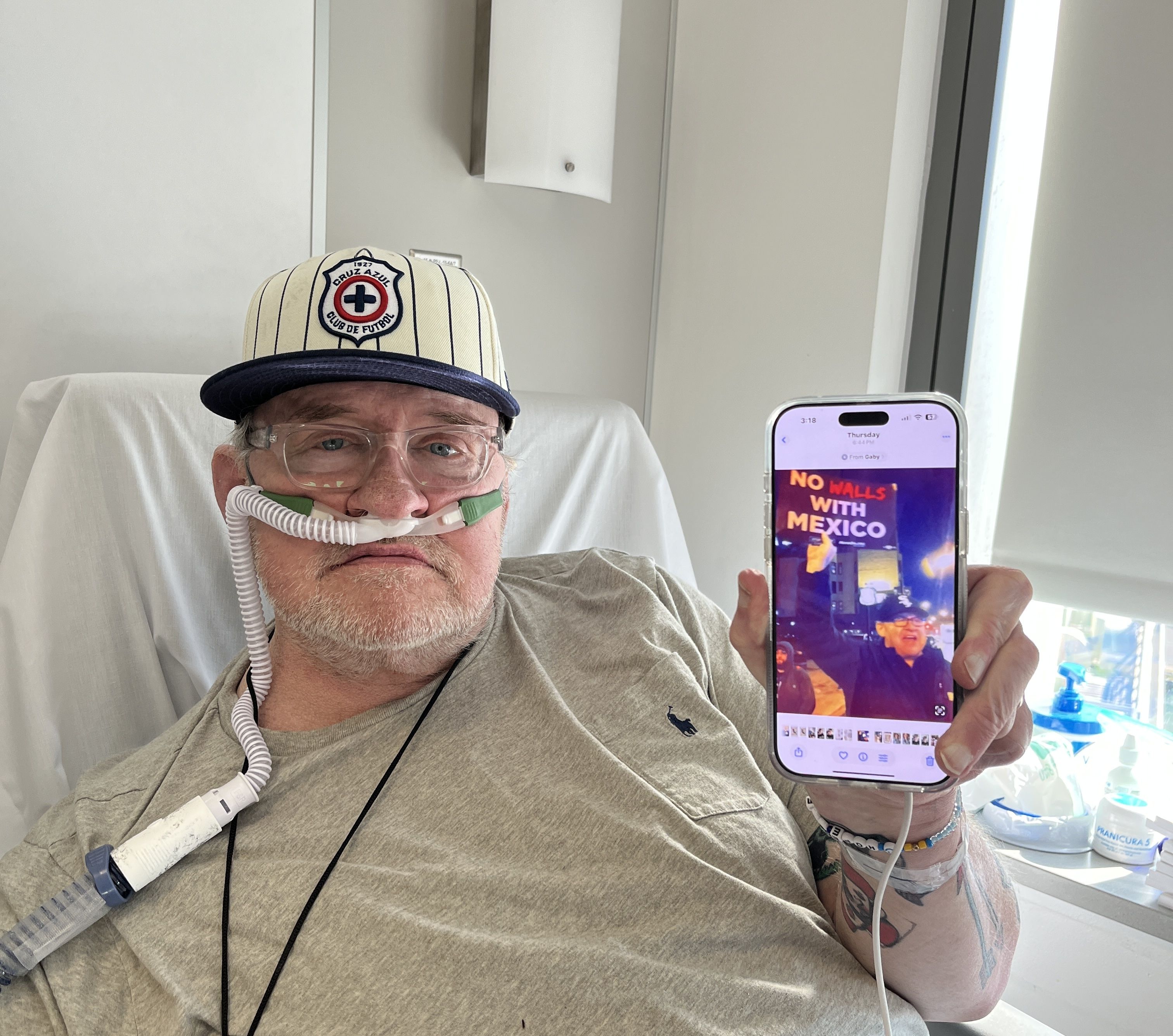 Man in a gray shirt and striped Cruz Azul cap with oxygen tubing holds a phone showing a protest sign reading "NO WALLS WITH MEXICO" in a bright room.