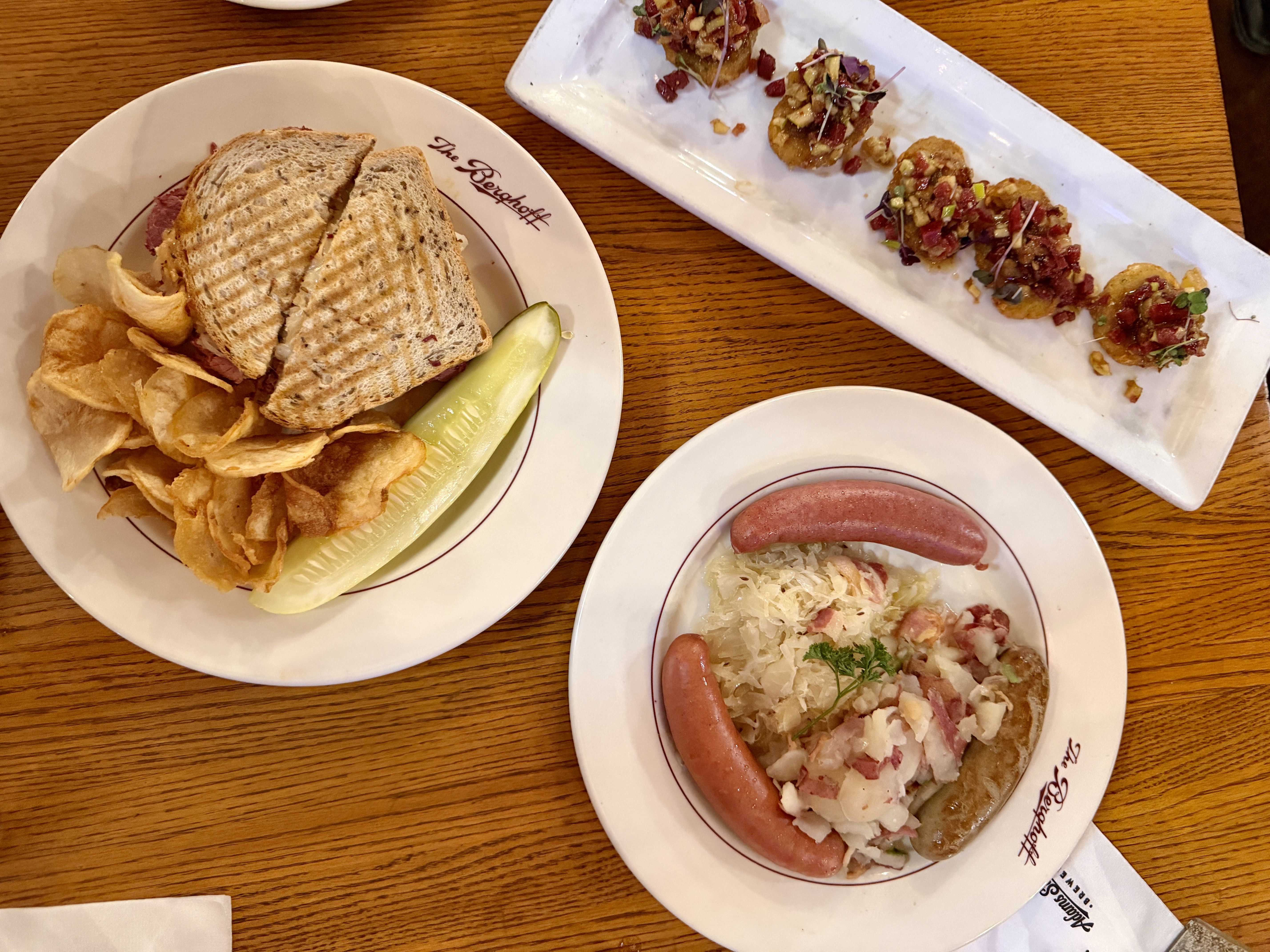 Three plates on a wooden table: a sandwich with chips and a pickle, a plate with two sausages, sauerkraut, and chunky potatoes, and a rectangular plate with five bite-sized appetizers topped with diced garnish.