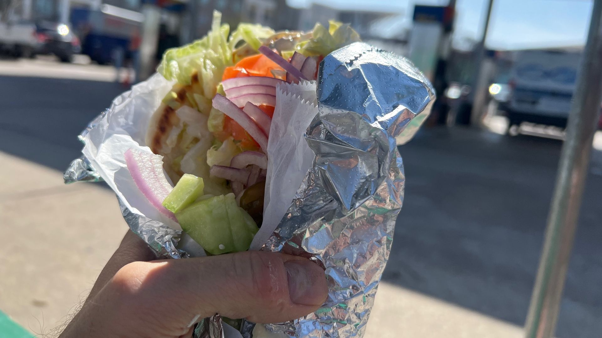 A hand holding a falafel wrap in tin foil with a gas station in the background.