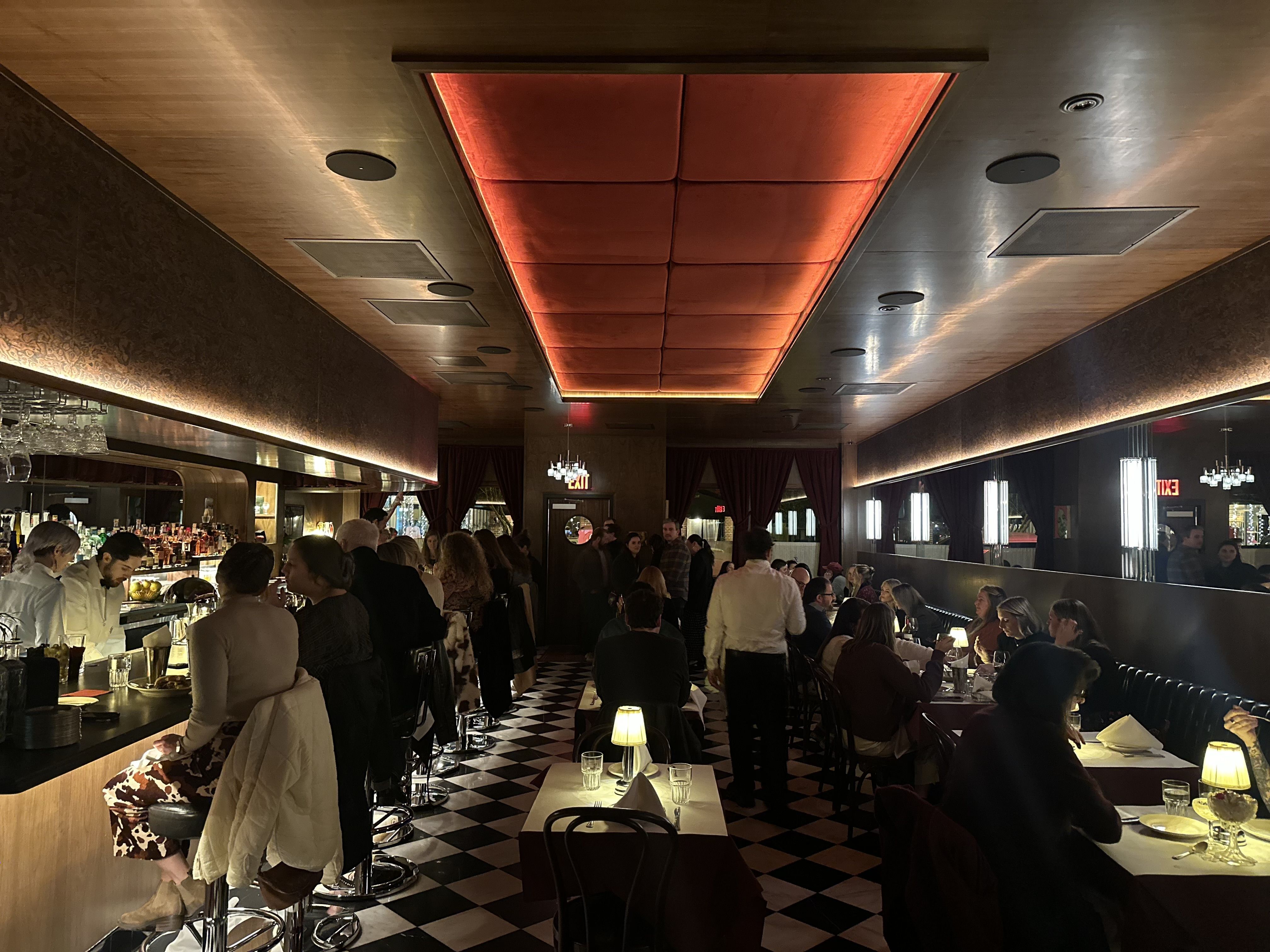 Dimly lit restaurant interior with black and white checkered floor, red ceiling panel, long bar on left with seated patrons, tables with lamps on right, and groups dining.