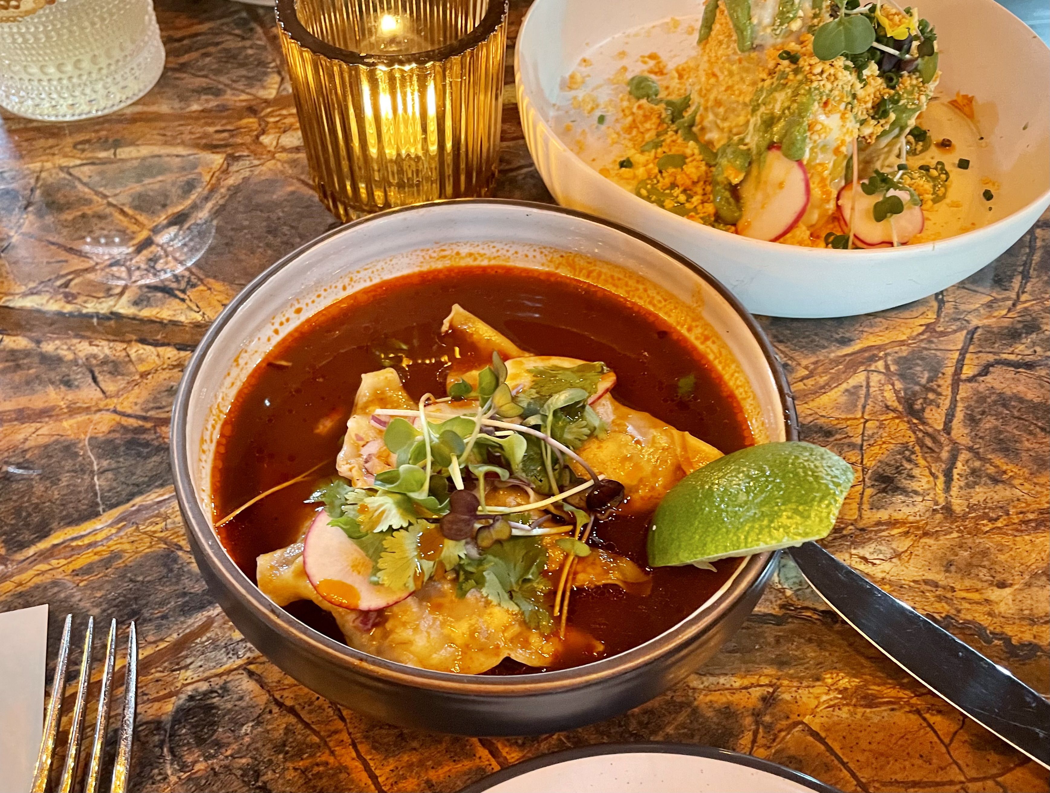 Two bowls on a marbled table: a dark red soup garnished with greens and radish, and a white bowl of crunchy salad with microgreens. An amber candle glows between them, with lime and utensils nearby.