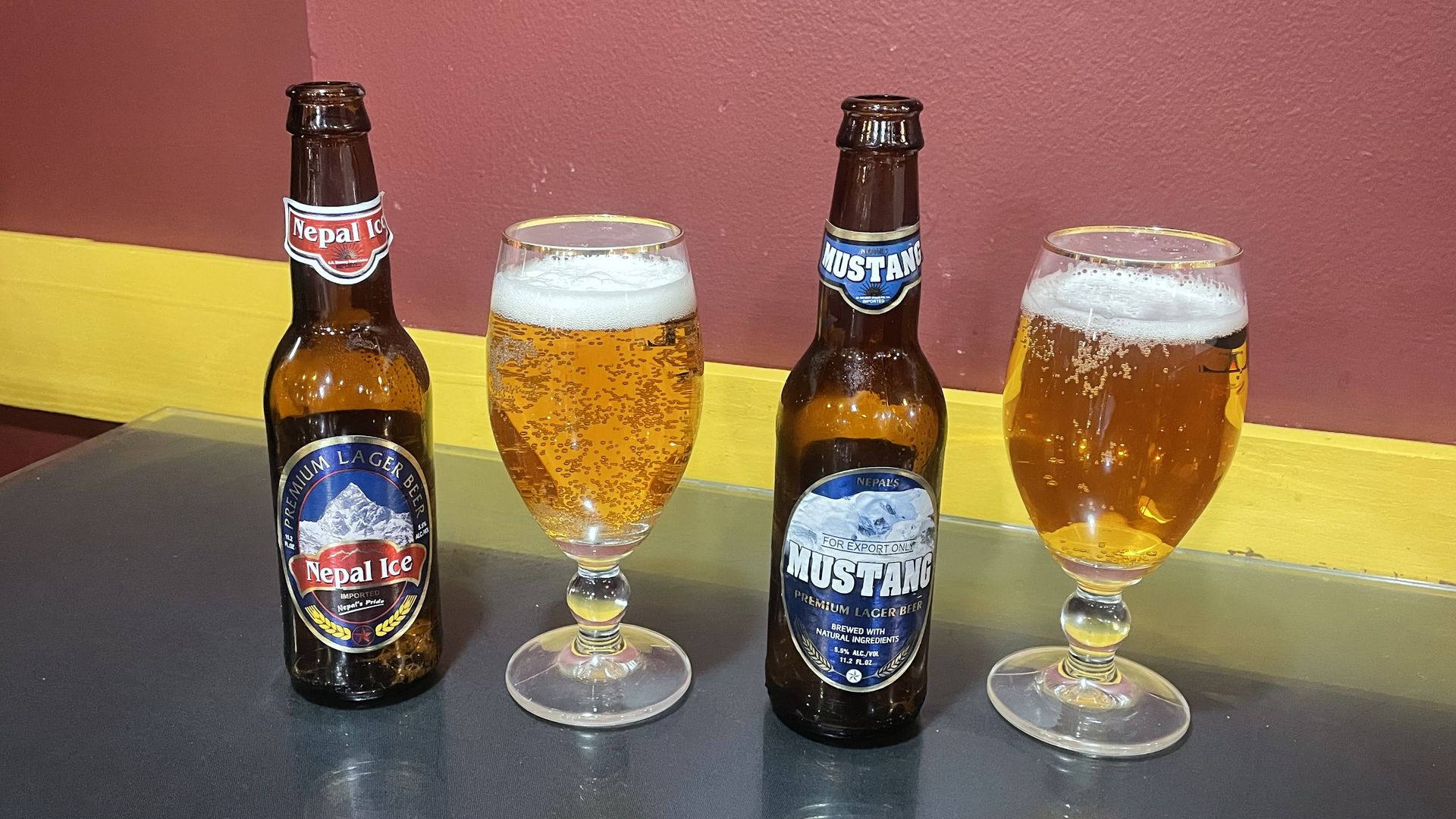Two beer bottles (Nepal Ice left, Mustang right) flank two stemmed glasses of amber beer with foamy heads, set on a dark table against a maroon wall with a yellow stripe.