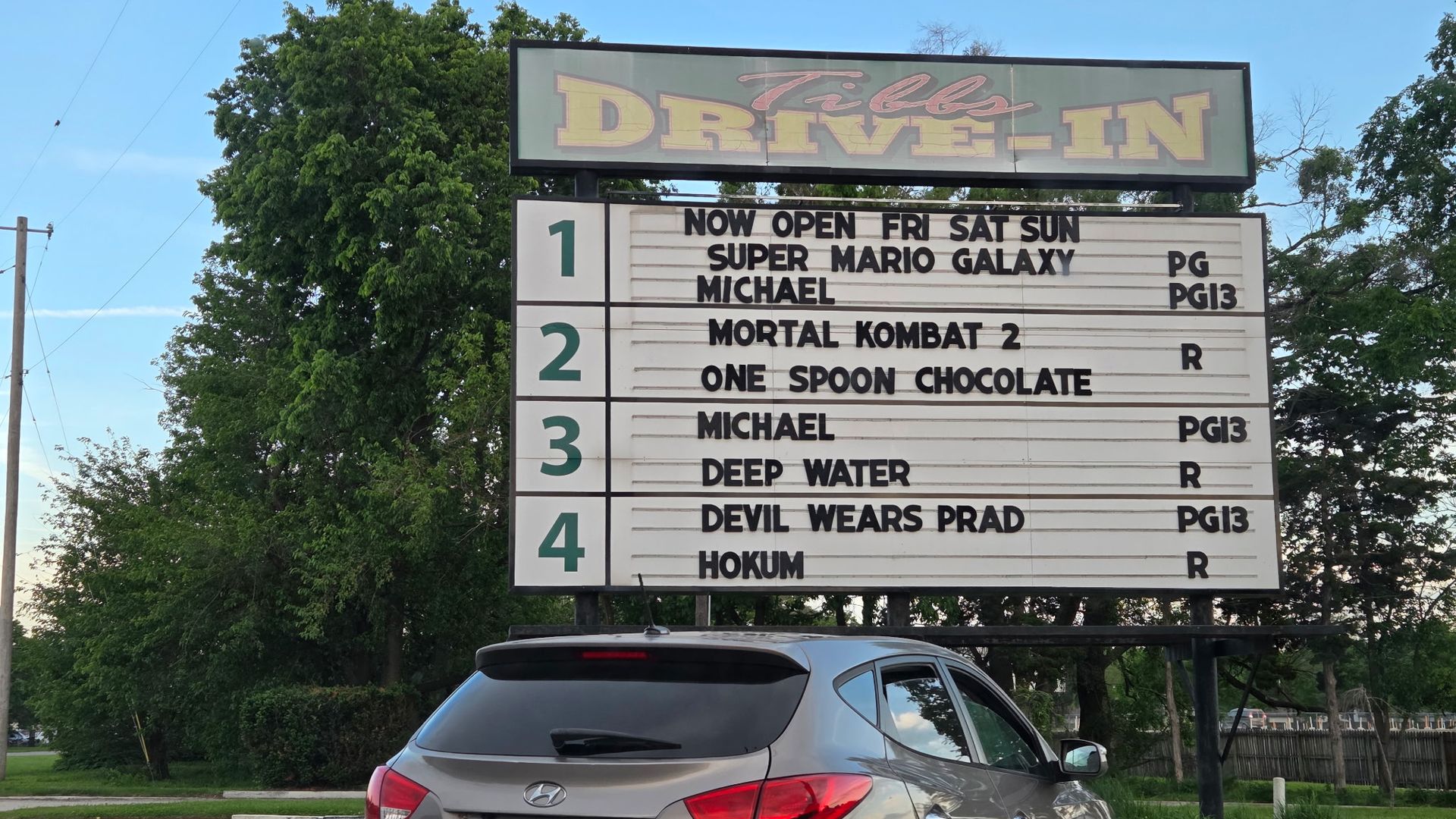 Drive-in theater marquee with a large green DRIVE-IN sign above a white board listing movie titles; a gray Hyundai SUV with Indiana plates parked in front, blue sky and trees behind.