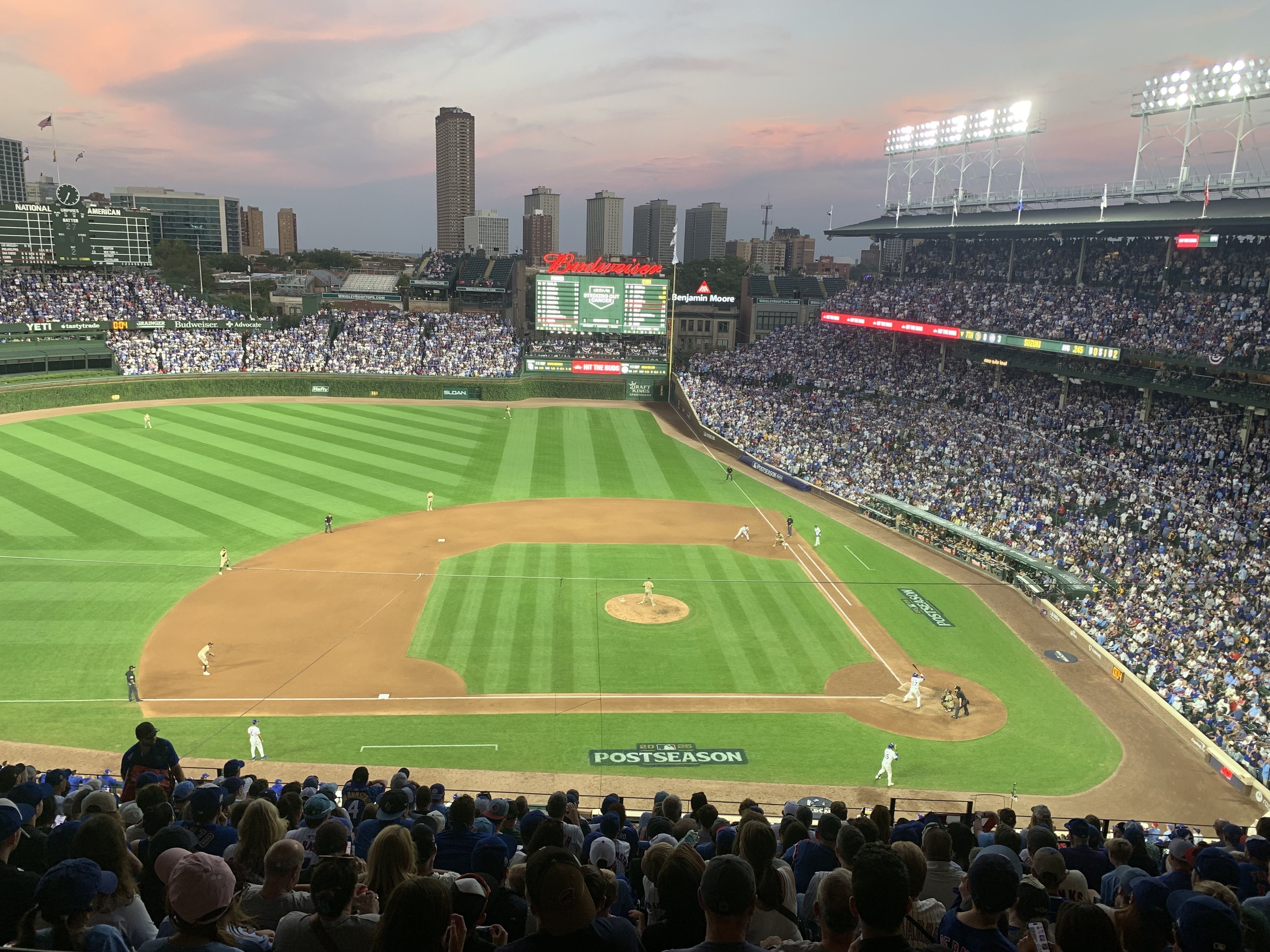 Crowded Wrigley Field during sunset with green field, players in white and beige uniforms, and a large scoreboard displaying postseason information, surrounded by city skyscrapers and pink sky.