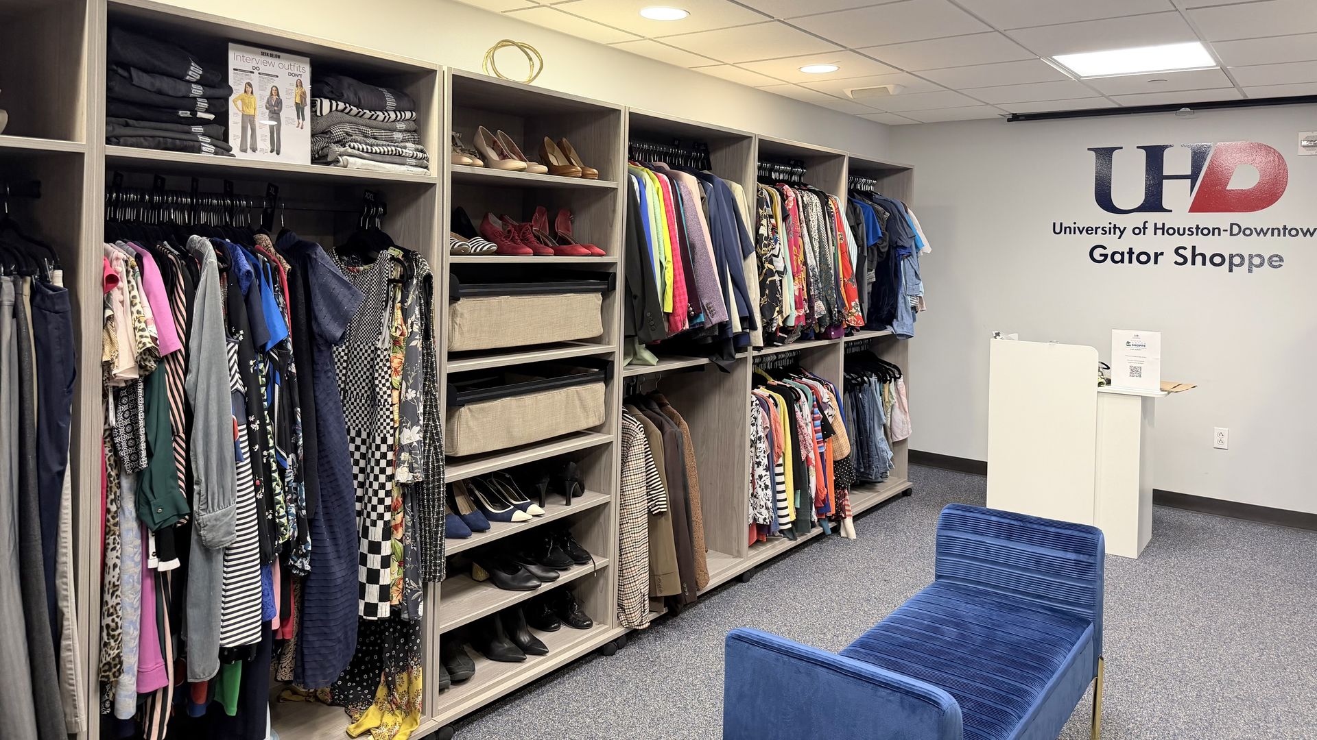 Interior of University of Houston-Downtown Gator Shoppe with racks of colorful clothes, shelves of shoes, and a blue cushioned bench on a carpeted floor.