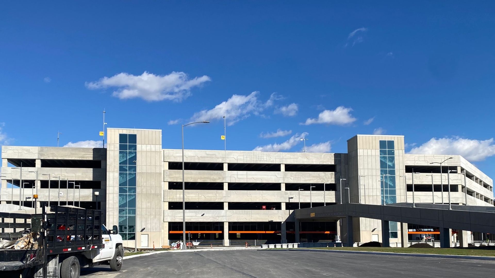 Wide view of a modern multi-level concrete parking structure with glass elevator towers under a blue sky with clouds and a white truck parked nearby.