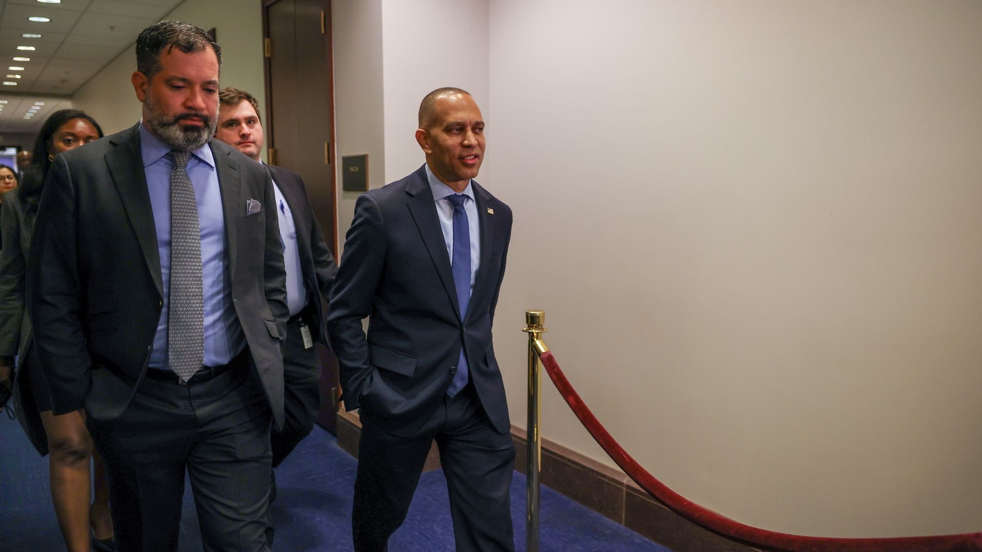 House Minority Leader Hakeem Jeffries wearing a blue suit and walking through a white hallway with retinue of aides.
