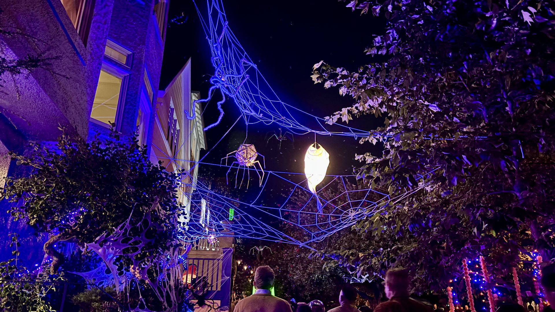 Night scene with blue neon spider-web lights crisscrossing above a street, a glowing lantern center, purple foliage, a building left, and people seen from behind.